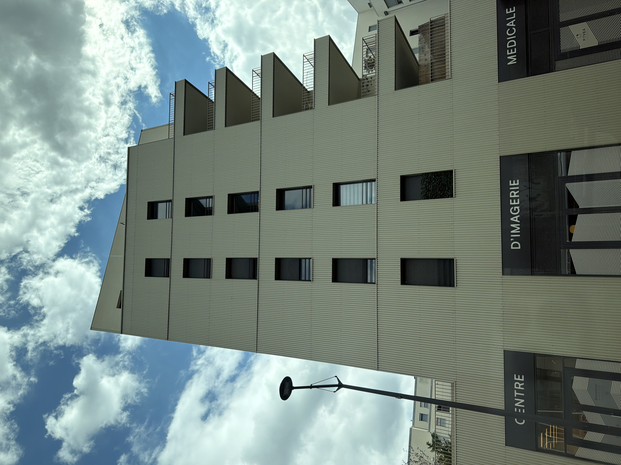 A modern, tall building with a light beige, corrugated facade stands against a blue sky with scattered white clouds. The building has multiple levels with dark-framed windows and balconies with railings. Street level signage indicates a "CENTRE," "D'IMAGERIE," and "MEDICALE" facility. A tall, dark lamppost is positioned to the left of the building. The scene is likely in Ivry-sur-Seine, France, as indicated by similar architectural styles and street names found in the area. There are no people visible in the image.