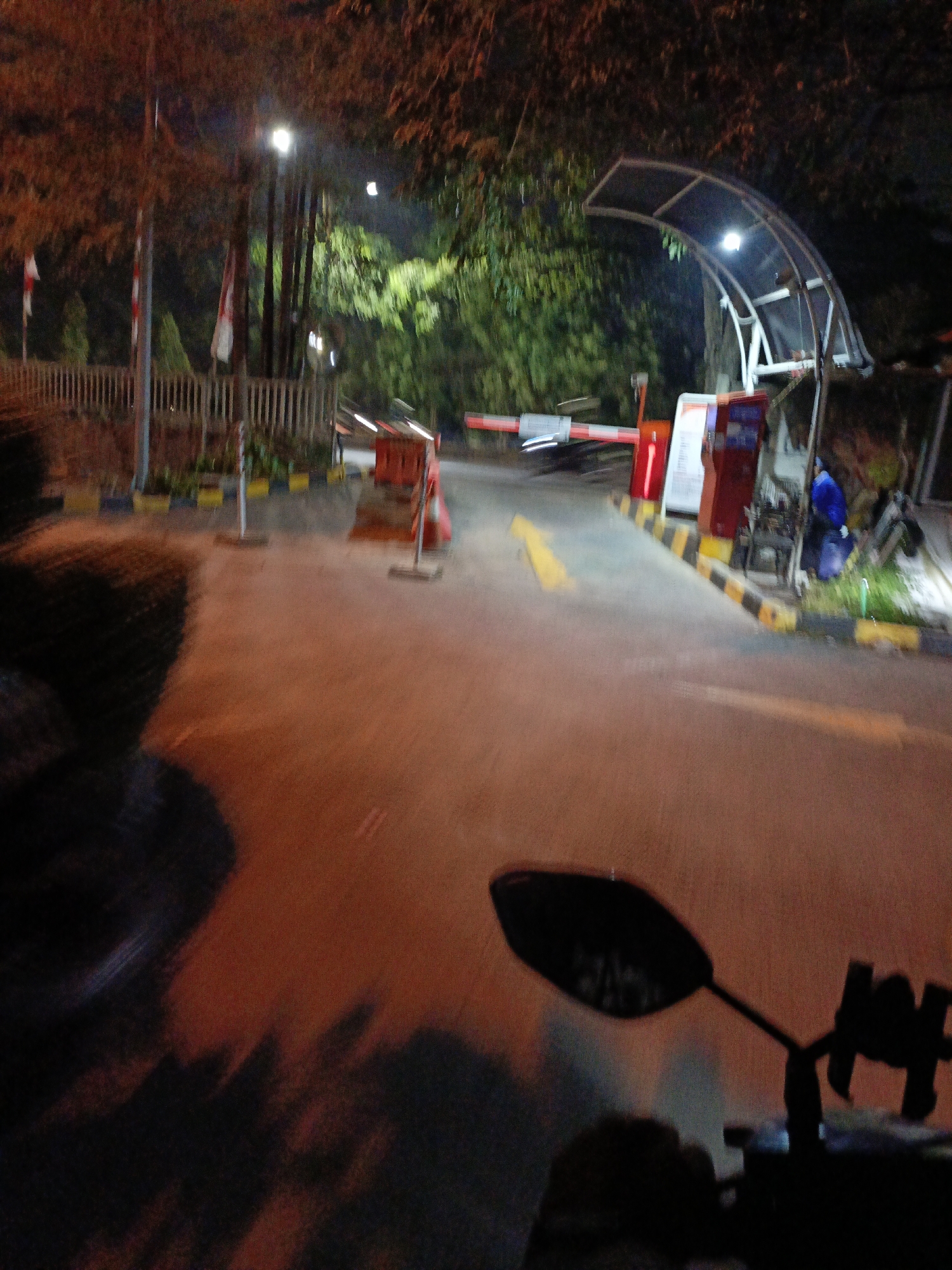 In Wangunharja, Indonesia, at night, a motorcycle approaches a manned checkpoint. The checkpoint features a red and white boom gate, two red payment terminals, and a white arched canopy. A person in a blue jacket is visible in the background, crouching near the terminals. The road surface is asphalt with yellow lane markings. The surrounding environment includes trees with illuminated leaves, a wooden fence, and a street light. The image is taken from the perspective of the motorcycle rider, with the motorcycle's rearview mirror and a portion of its handlebar visible in the foreground.