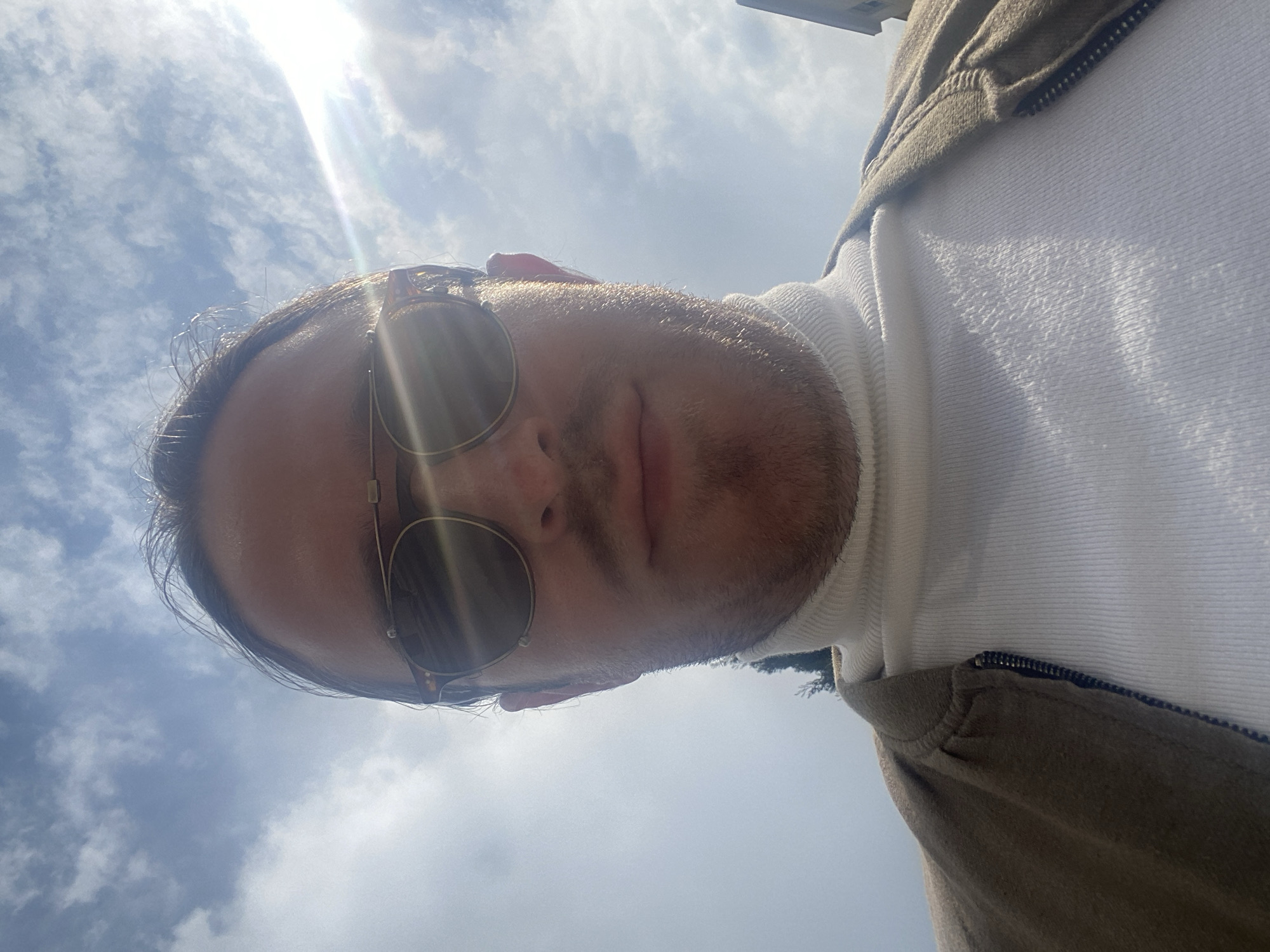 A male individual is depicted in a low-angle selfie, positioned against a bright, cloudy sky in Budva, Montenegro. The person wears dark, round sunglasses with a gold-colored metal bridge and patterned frames, which reflect a prominent sun flare originating from the upper right of the frame. A white, ribbed turtleneck sweater is visible, covered by a partially unzipped light brown or tan jacket with a quilted or textured appearance on the shoulder. Facial stubble and a mustache are present. The background consists primarily of a bright blue sky with scattered white cumulus clouds. The sun, located in the upper right, casts strong backlighting on the subject. A faint dark, slender vertical object, possibly a tree or distant structure, is visible on the left side, behind the subject's left shoulder. A light-colored building structure is partially visible on the far right edge of the frame. The individual appears to be looking directly into the camera.