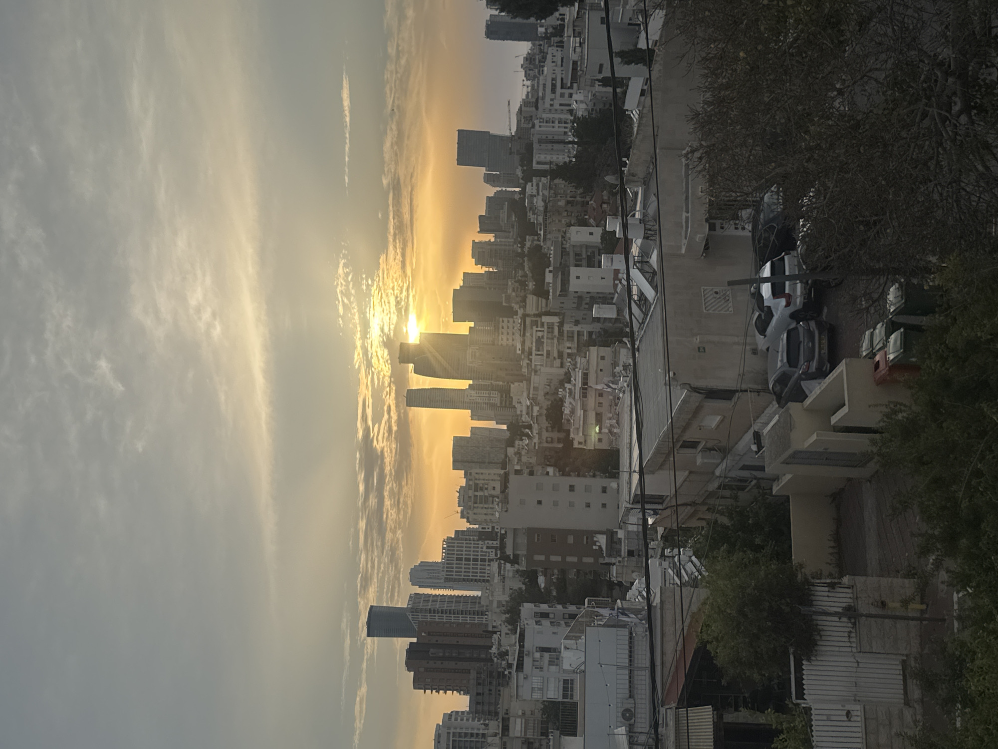 A cityscape is captured during either sunrise or sunset, with the low sun radiating a brilliant golden light across the sky on the left side of the frame. Wispy clouds are illuminated by the sun, creating streaks of bright orange and yellow against a lighter blue-grey sky.
The prominent feature is the urban sprawl of buildings, stretching into the distance. Numerous multi-story buildings, including several taller high-rises, are silhouetted against the bright sky, showcasing modern and older architectural styles. The buildings in the midground and background appear denser and more clustered, typical of a bustling city.
In the foreground and right side of the image, residential or commercial buildings with visible balconies, windows, and utility elements like air conditioning units are present. The immediate foreground shows a street scene with several parked cars. Dark, dense foliage from trees, possibly olive trees or similar species, obscures parts of the foreground and cast shadows, adding a natural element to the urban environment. Utility poles and wires crisscross the upper right portion of the scene.
There are no people visible. The time of day is clearly either early morning or late evening, characterized by the dramatic light and long shadows. Given the explicitly provided location context, this is a scene from Ramat Gan, Israel. No discernible text is visible on any signs or buildings. The overall impression is one of urban life at a specific, beautiful transitional moment of the day.