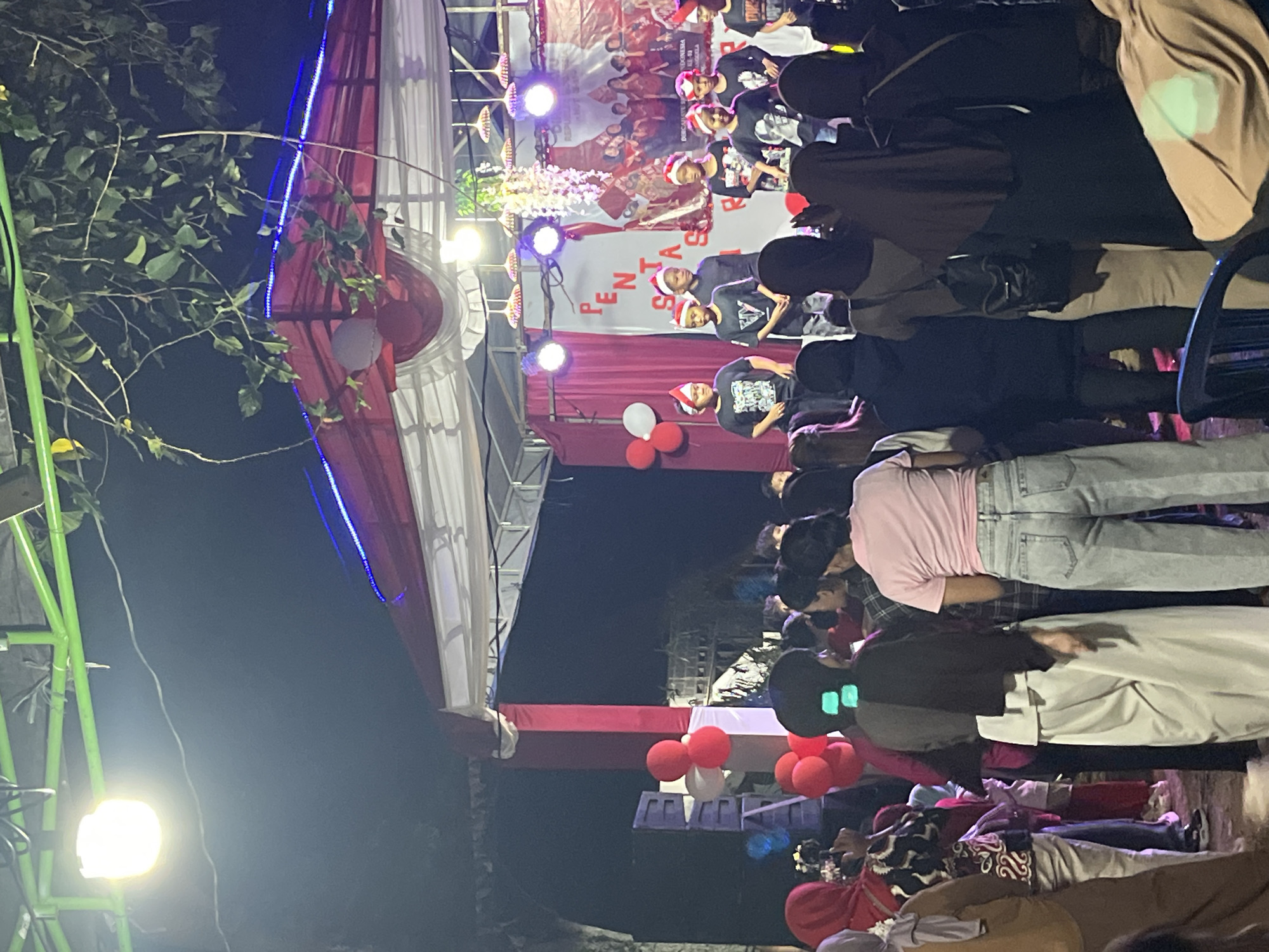 The image depicts an outdoor evening event in Tajurhalang, Indonesia, likely a local festival or performance. A stage is set up, adorned with red and white fabric and illuminated by blue and white lights. Several children, wearing red and white headbands, are performing on stage, dressed in black t-shirts. A banner behind them displays text that is partially visible. A crowd of spectators, many wearing headscarves, are gathered in front of the stage, watching the performance. Balloons in red and white are visible, adding to the festive atmosphere. The environment appears to be a public space, with trees and outdoor lighting visible in the background.