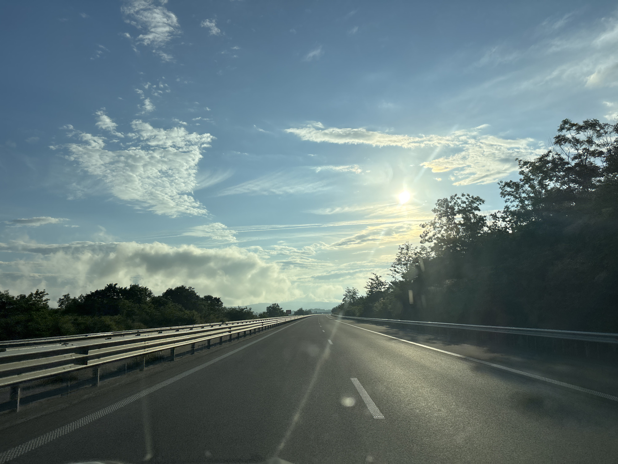 The image depicts a road, likely a highway, stretching towards a distant horizon under a partly cloudy sky. The sun is visible and shining brightly, casting a warm glow and creating lens flares on the road surface. On either side of the road, there are guardrails and dense green trees. The road itself is asphalt with clear white lane markings. The sky features a mix of scattered white clouds and wispy cirrus clouds against a pale blue backdrop. The scene appears to be captured during the daytime, possibly late afternoon or early morning due to the angle of the sun. The location of Yazdach, Bulgaria is not identifiable from the visual information provided in the image.