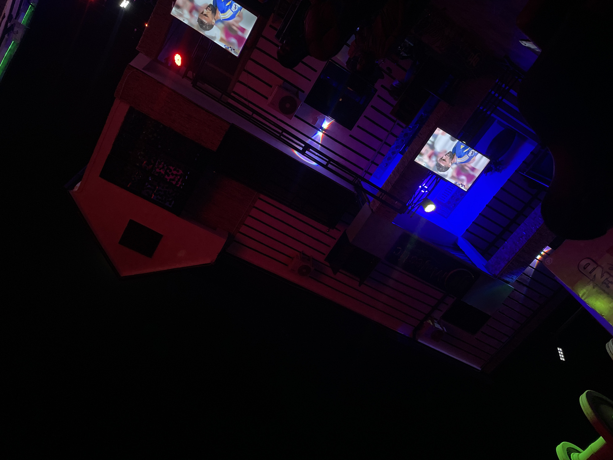 The image, taken at night in Tumfure, Nigeria, shows a building with multiple levels and exposed roof beams. Two televisions are visible, each displaying a soccer game. The building is lit with red, blue, and green lighting. A person can be seen in the top of the frame. A sign and other objects are present in the bottom right.