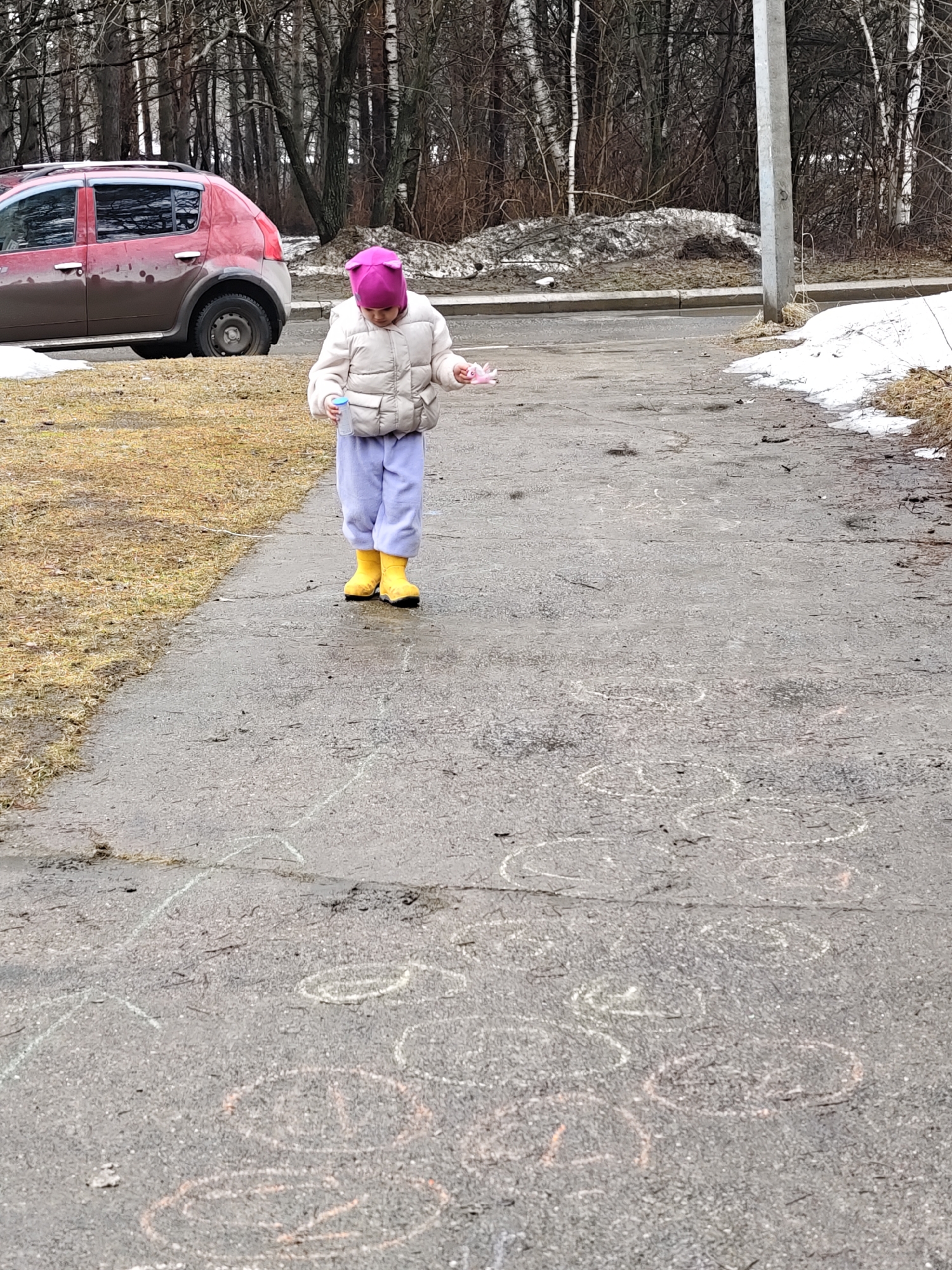 A young child in a pink hat, light-colored jacket, light purple pants, and yellow boots stands on a gray asphalt surface. The child appears to be holding a small bottle in one hand and a pink object in the other. The ground is marked with chalk drawings. A red car is partially visible in the background. Patches of grass and trees are also in the background, along with remnants of snow. The location appears to be Chaikovsky, Russia, based on the image context.
