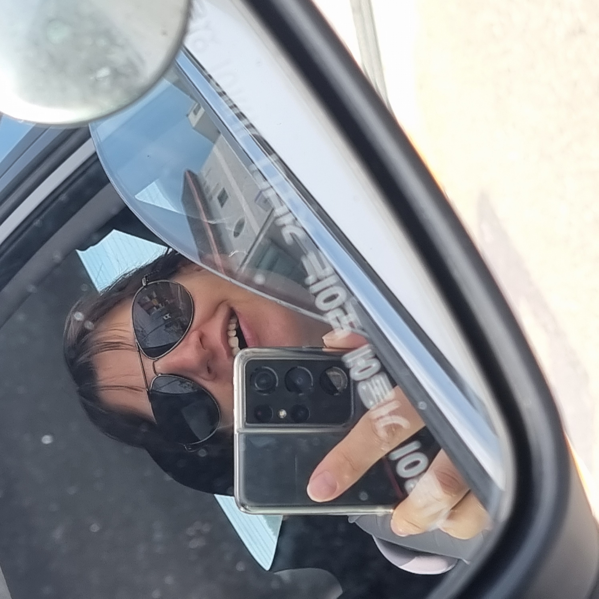 A woman wearing sunglasses and a light-colored jacket is reflected in the side-view mirror of a car, taking a selfie with her phone. She is in the driver's seat, and her phone is held up, capturing her image and a portion of the car's interior. The reflection in the mirror shows the surrounding environment, including buildings and the sky, indicating she is likely in an urban or suburban area. Text on the phone case and a sticker on the dashboard are partially visible. The scene appears to be outdoors, with natural light.