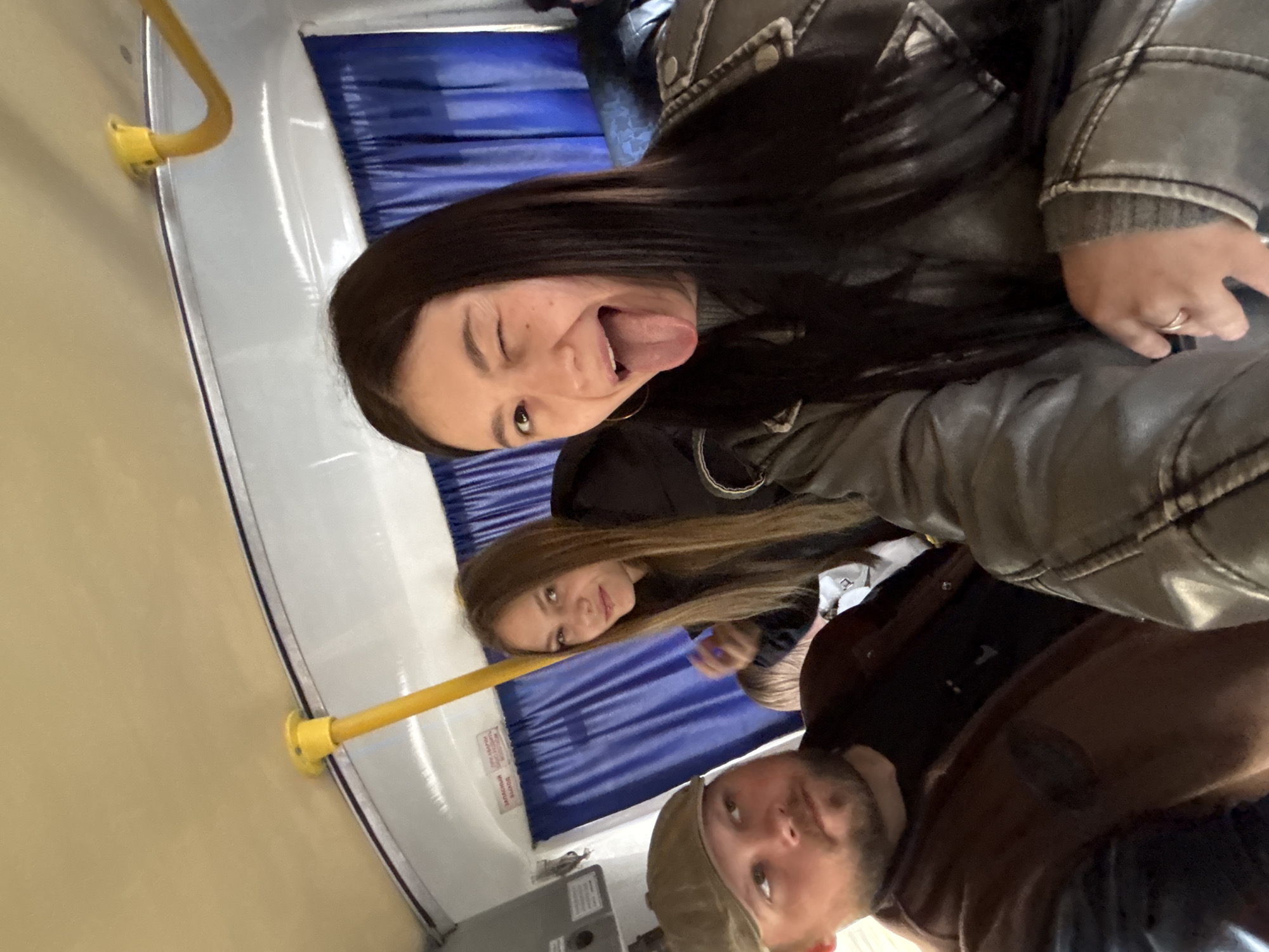 The image shows a selfie taken indoors in what appears to be a public transport vehicle, potentially in the city of Chaikovsky, Russia. Three individuals are visible. The person closest to the camera is a woman with long, dark hair wearing a grey leather jacket. She is making a face with one eye closed and her tongue sticking out. Behind her, another woman with long brown hair is looking at the camera. At the bottom of the frame, a man with a beard and a baseball cap is also looking upwards. The background reveals blue padded seating with white trim, yellow handrails, and potentially a wall-mounted electrical box. The lighting seems artificial, and the image is slightly angled, indicating it was likely a handheld shot.