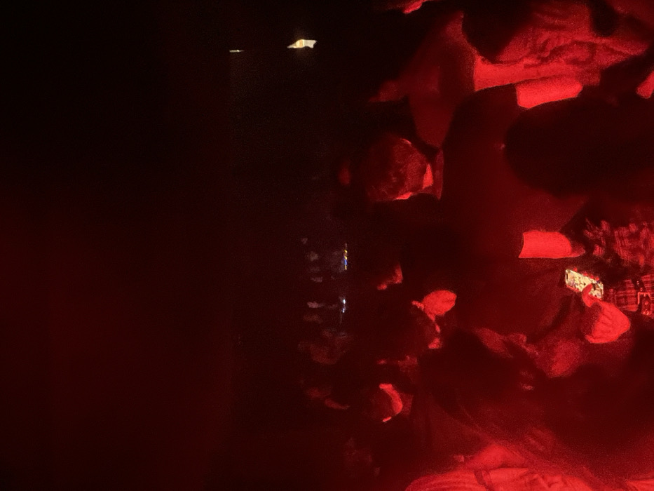 The image depicts an indoor scene within a crowded nightclub in Barcelona, Spain, consistent with venues such as Sala Apolo, illuminated predominantly by deep red ambient lighting. The upper portion of the frame is entirely obscured by darkness. In the mid-ground, a person with dark hair is observed looking down at an illuminated smartphone held in their hands. The phone's screen casts a bright, white-blue light upwards onto their face and chest, revealing a patterned garment. This individual is surrounded by numerous other patrons, visible primarily as dark silhouettes and partially red-lit forms; their faces and specific details are largely indistinguishable due to the low, monochromatic light. A dense crowd of people is present throughout the mid-ground and extends into the background, standing in close proximity. In the far background, additional indistinct figures are visible, some appearing to be on a slightly elevated platform or stage area. The overall scene indicates a high-density social gathering or music event.