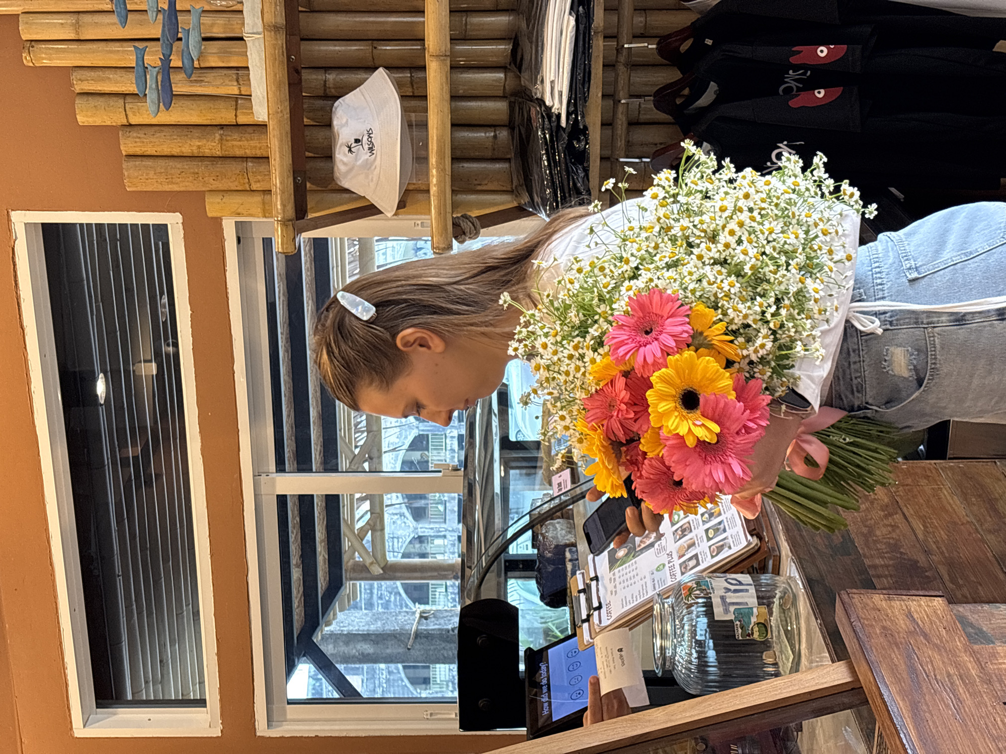 A person stands indoors near a wooden counter in Phuket City Municipality, Thailand. A bouquet of pink, orange, and white flowers is positioned prominently in the foreground, held by the person, who is wearing a white top and denim pants. The person is looking down at a menu on the counter, next to an electronic device with an evaluation question displayed. A jar sits nearby. The background includes a window with a view of the cityscape, decorative bamboo, and clothing hanging.