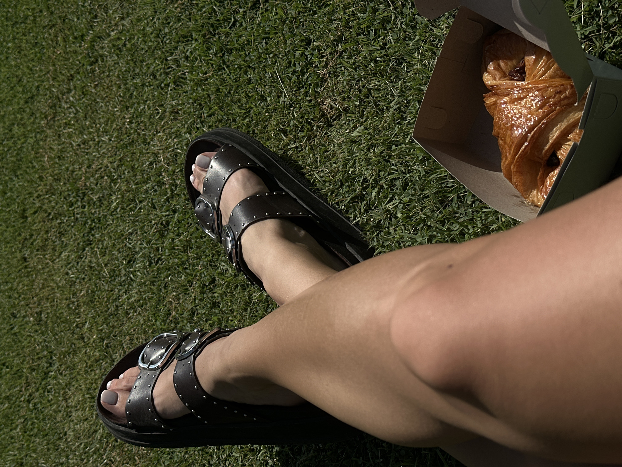 The image depicts a partial view of a person seated on green grass outdoors. The person's legs are visible, with one leg crossed over the other. They are wearing black sandals with studded straps and silver buckles. Next to their legs, on the grass, is an open box containing a pastry, likely a chocolate croissant. The specific location in Unknown, Indonesia is not identifiable based on the image.