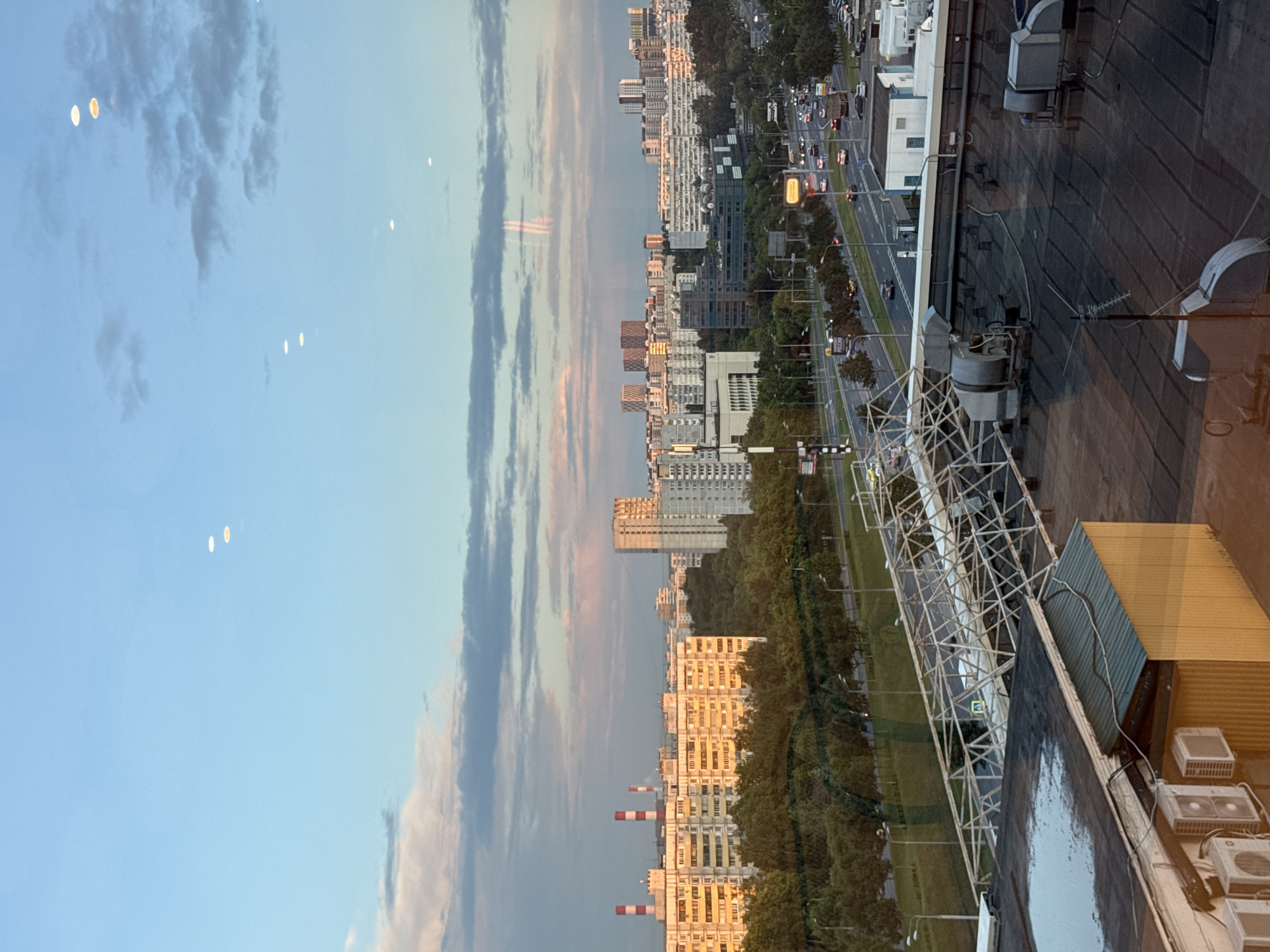 The image, taken from an elevated perspective likely from a building, depicts a city scene in Moscow, Russia, during what appears to be late afternoon or early evening, given the warm, golden light reflecting off some of the buildings. The viewpoint is looking across a street lined with trees, towards a row of tall, multi-story residential buildings. These buildings are a mix of architectural styles, with some appearing older with balconies and others more modern. Two prominent industrial chimneys with red and white bands are visible amongst the buildings.
The foreground shows the dark, flat roof of the building from which the photo is taken, equipped with various rooftop units, possibly air conditioning or ventilation systems, and a large, angled metal structure resembling scaffolding or a support framework. To the right, a portion of a yellow structure is visible.
Beyond the buildings, a busy street is visible with cars driving and streetlights illuminating the road. Trees form a green buffer between the street and the residential buildings. The sky is a soft blue, with wispy clouds that are tinged with pink and orange hues, suggesting the setting sun. Several bright, circular or oval reflections are scattered across the sky, likely caused by lights or lenses.
No people are explicitly visible in the image. The overall mood is one of urban life transitioning into evening, with a sense of scale provided by the tall buildings and the expanse of the sky.