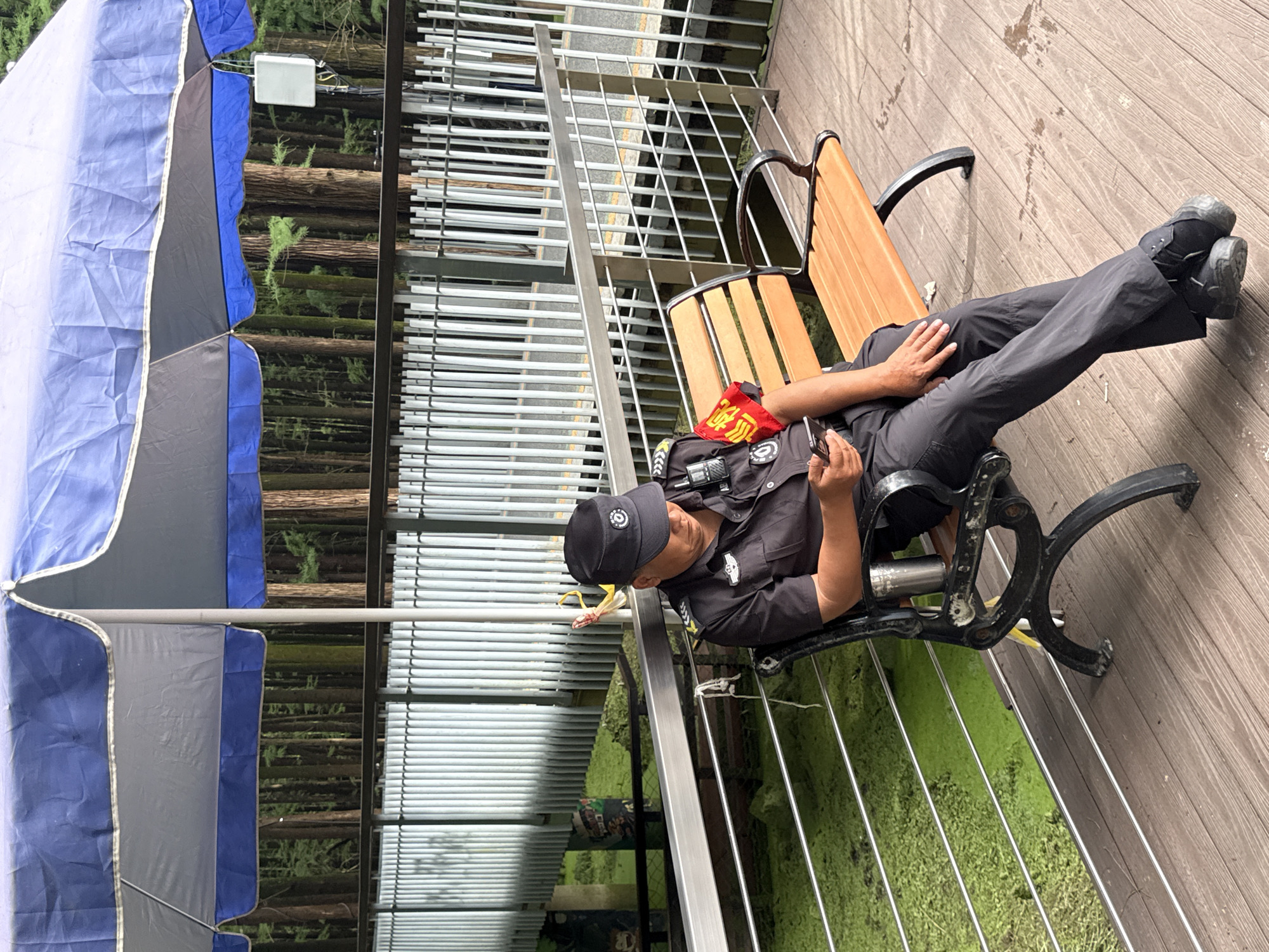A uniformed security guard, wearing a black cap and dark uniform with a red armband, sits on a wooden bench under a large blue and gray umbrella. The guard is looking down at a smartphone in their hands. A silver thermos is attached to the side of the bench. The bench is situated on a wooden deck. Beyond the deck is a railing and then a body of green, algae-covered water. In the background, a dense forest of tall trees is visible, with a white fence running horizontally across the middle ground. A white box with wires is attached to a pole to the right of the trees. The scene appears to be an outdoor public area, possibly a park or a recreational spot within Lin'an District, China, given the context provided.