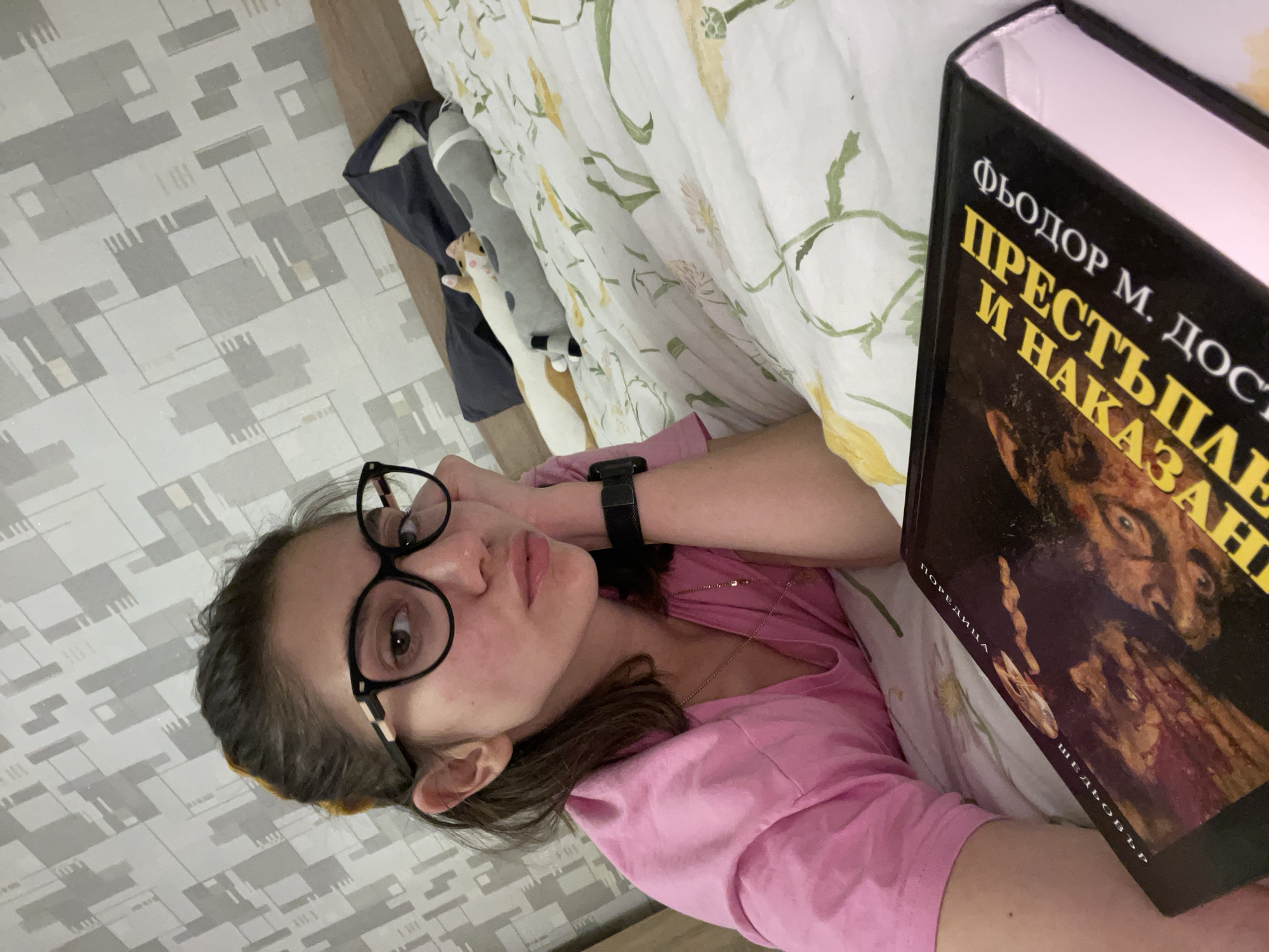 A young woman wearing glasses and a pink top is lying in bed, reading a book. The book is in Russian. The title of the book is "Crime and Punishment" by Fyodor Dostoevsky. The bed has a white floral patterned comforter. The woman is wearing a watch on her left wrist. A stuffed animal is visible near the head of the bed. The room has patterned wallpaper. The location is likely an apartment. Without further visual or contextual clues, it's impossible to determine if the location is in the city of Sofia, Bulgaria.