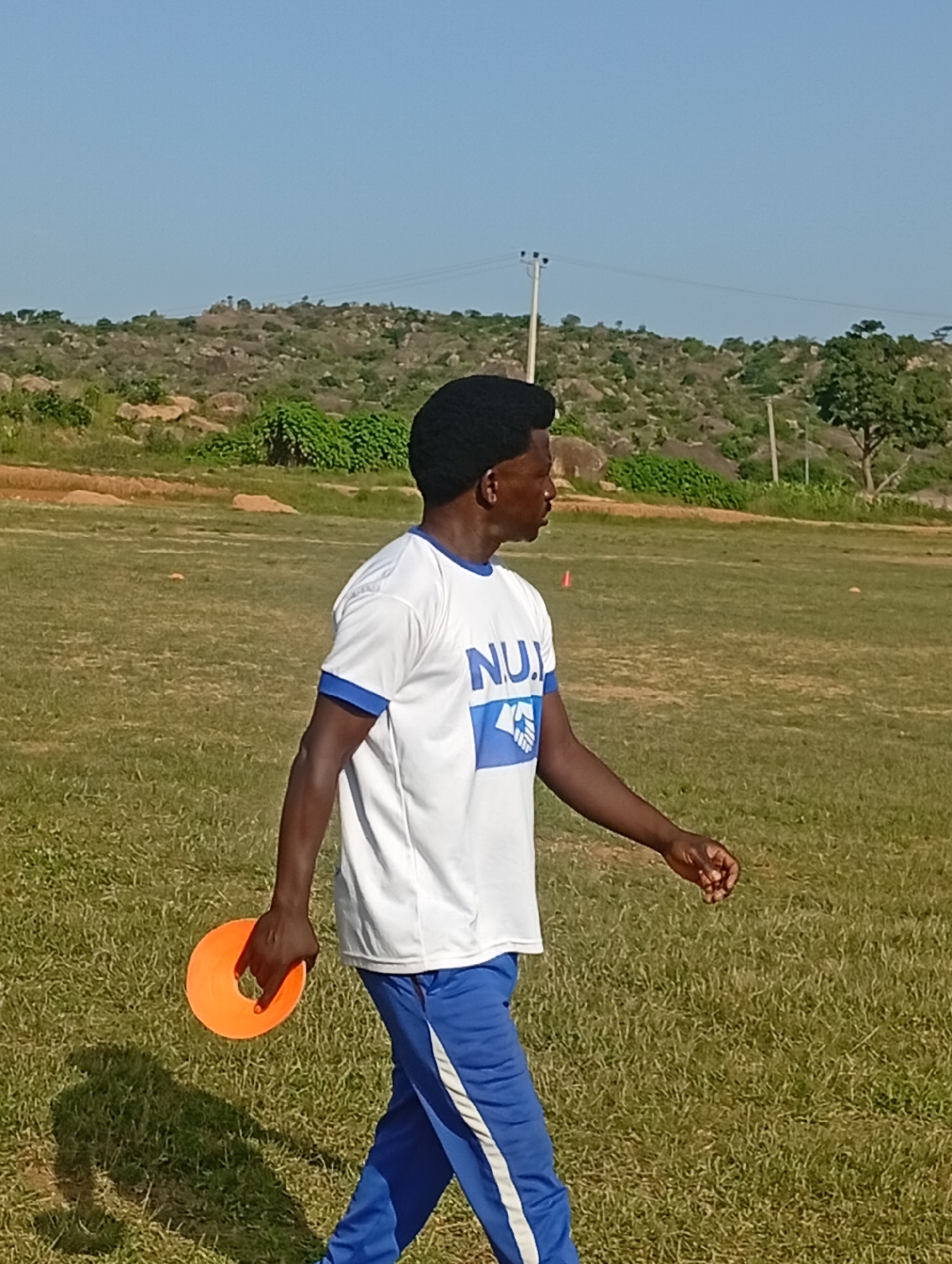 A medium shot captures a Black male from the chest down, walking from left to right across a grassy field. He is wearing a white t-shirt with blue trim around the collar and sleeves, and blue athletic pants with a white stripe down the side. The t-shirt has a blue logo with white lettering that reads "NUJ". He is holding an orange disc-shaped object in his right hand. The background shows a dry, grassy field with scattered rocks and shrubs, and a gentle slope in the distance under a clear blue sky. There are a few utility poles visible in the background. The scene appears to be an outdoor athletic field in a rural or undeveloped area.