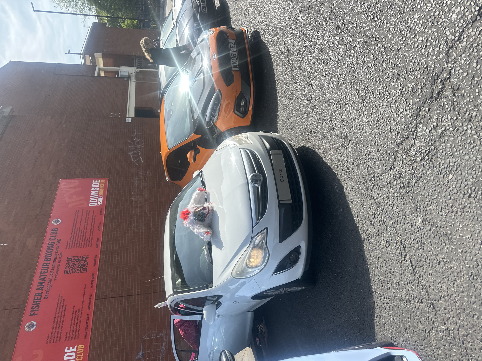 Here's a detailed description of the scene:
**Setting:** An outdoor street scene in London, United Kingdom. The weather is sunny, as indicated by the harsh shadows and bright sunlight reflecting off the vehicles.
**Objects:**
* **Vehicles:** Three cars are visible. A white Vauxhall Corsa is in the foreground, followed by an orange Ford Fiesta, and a dark-colored vehicle (possibly a black car) further back. The Ford Fiesta's registration plate is partially visible, showing "MK08 FEZ". The dark car's numberplate visible is "K20 LBP"
* **Building:** A brick building stands on the left side of the frame. A large red banner attached to the building advertises the "Fisher Amateur Boxing Club" and "Downside Fisher Youth Club."
* **Road:** The street is paved with dark asphalt.
* **Decorations:** A furry, possibly festive, decoration sits on the hood of the white Corsa.
**People:**
* An adult is standing in front of the orange Ford Fiesta.
**Actions/Interactions:**
* The adult is either leaning on or working on the Ford Fiesta.
* The white Vauxhall Corsa has its driver-side door open, partially revealing the interior.
**Environment:**
* The background includes elements of an urban environment, such as street furniture (street light) and parts of other buildings.
* Graffiti is visible on the brick building behind the cars.