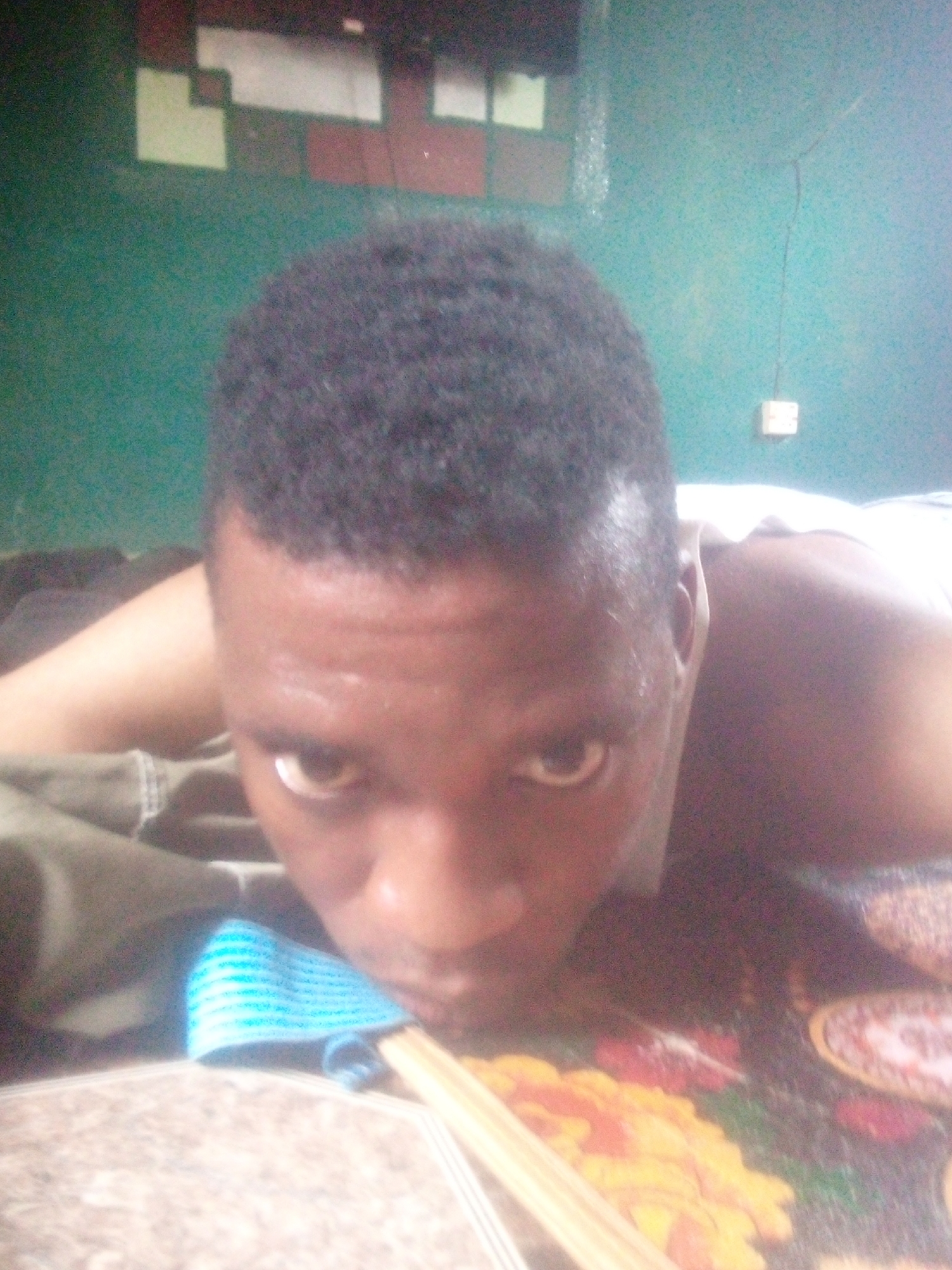 An eye-level, close-up shot depicts a person, likely an adult male with dark skin and short, curly dark hair, lying prone and looking directly into the camera. His face is central in the frame, with both eyes wide open and a neutral expression.
The person's upper body, including his shoulders and arms, rests on a surface covered by a patterned fabric, possibly a bed sheet or blanket. This fabric features a vibrant, somewhat blurred floral or abstract design with prominent yellow and red hues. To the left of the person, a section of dark, possibly olive green, fabric is visible. In the bottom-left foreground, a distinct blue and white striped fabric, resembling a towel or small blanket, is present. At the very bottom edge, several thin, light-colored, linear objects, possibly wooden slats or part of a bed frame, are partially visible.
The background wall is painted in a muted teal or greenish-blue color. Towards the top of the wall, an arrangement of geometric shapes – squares and rectangles in white, light green, and reddish-brown – suggests painted decor or wall art. A white electrical outlet with a dark wire running upwards from it is clearly visible on the right side of the wall.
The overall lighting is somewhat dim and diffused, indicative of an indoor setting, possibly a bedroom or living space. The scene suggests a relaxed and informal moment. No text is visible in the image.