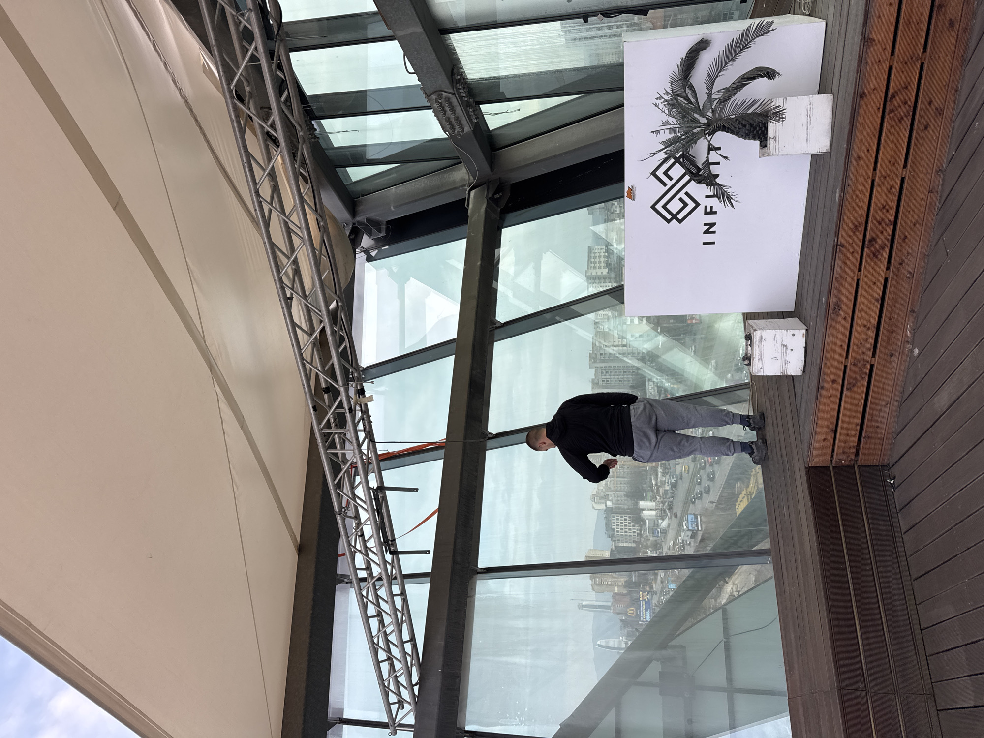 The image depicts an indoor/outdoor observation deck with a partial roof structure. A man stands on a wooden deck, looking down at the city visible through the glass panels. A white sign with a palm tree and the word "INFINITI" in a stylized font is positioned near the edge of the deck. A metal truss structure is visible, supporting part of the roof. Through the glass, a cityscape can be seen, identified as Sofia, Bulgaria, by the distinctive urban layout. The weather appears overcast.