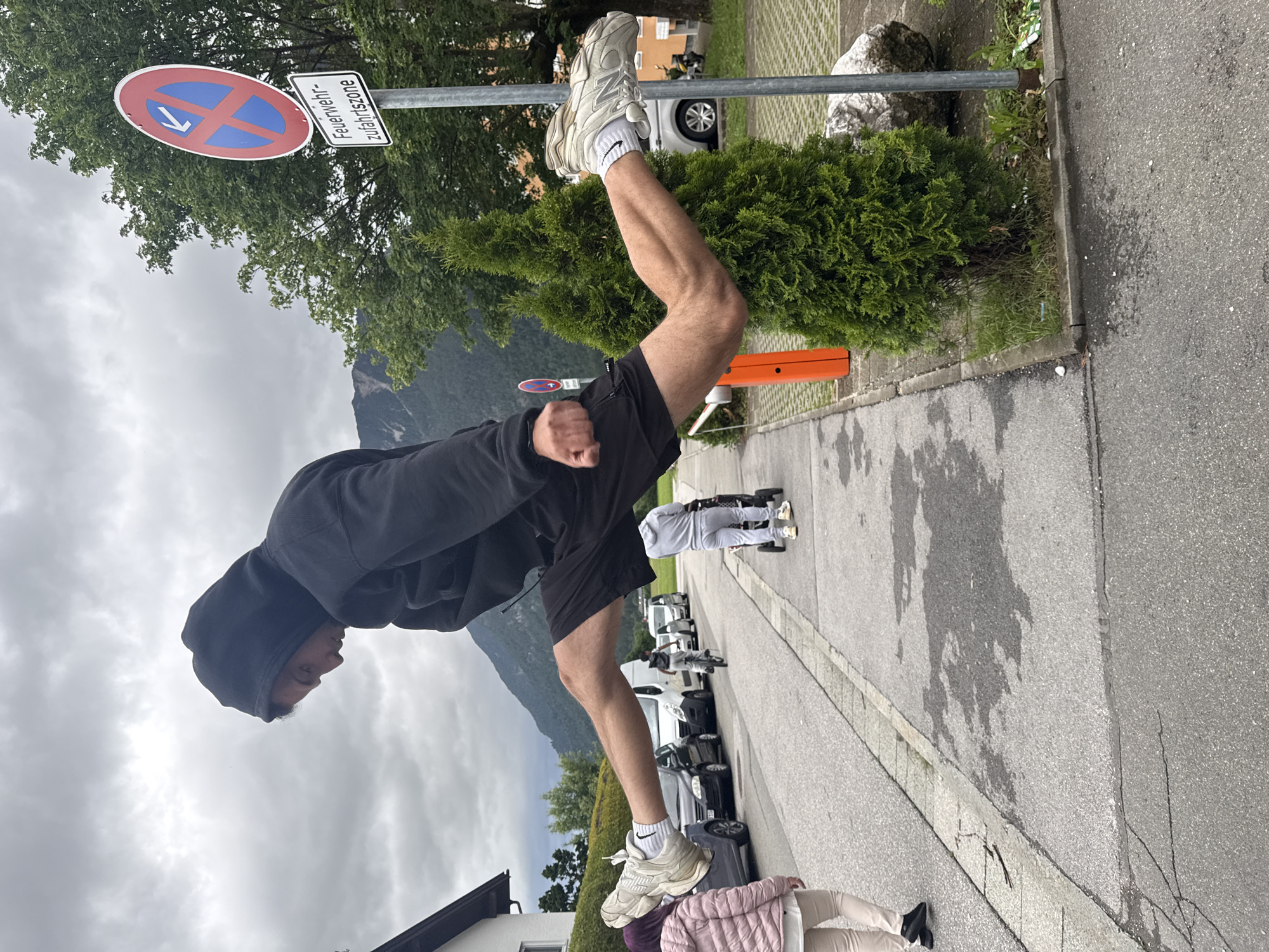 In Garmisch-Partenkirchen, Germany, a young male wearing a black hoodie and black shorts is captured mid-air performing a split jump on a cloudy day. He is positioned horizontally in the frame, with his legs extended in opposite directions. His left leg is forward and his right leg is backward. He is wearing white athletic shoes and white socks. In the background, a street is lined with parked cars and houses. A young person in a pink jacket and beige pants walks on the sidewalk to the left. Further down the street, another person rides a bicycle, and a third person pushes a stroller. A traffic sign indicating a fire department access zone is visible on the right, along with a small evergreen tree and a large rock. The sky is overcast with mountains in the distance.