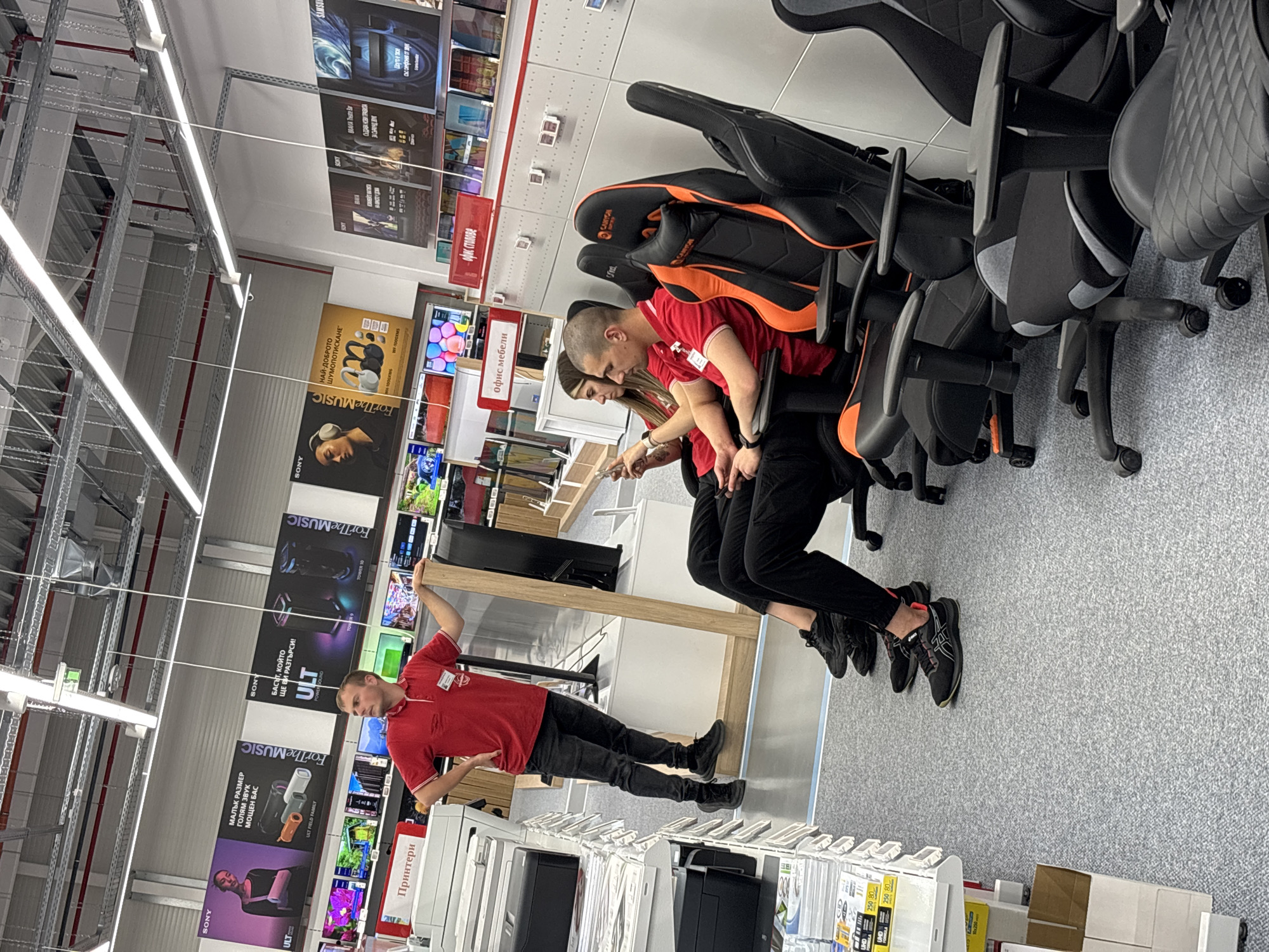 The photograph shows the interior of a retail electronics store, likely in Lovech, Bulgaria given the Cyrillic text in the background. A male employee wearing a red shirt and black pants is standing near a shelf unit of printers and pointing his arm towards a row of televisions mounted on the wall behind him. Two other employees, one male and one female, are seated in gaming chairs of varying black and orange colors, appearing to be focused on a mobile phone. The chairs are arranged in a line, suggesting a display. The flooring is a gray carpet. The ceiling has industrial lighting fixtures. Various promotional posters for audio and visual equipment, including Sony products, are displayed on the walls.