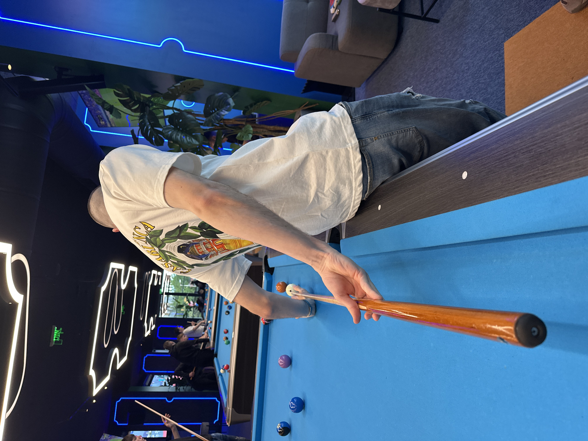 A person is playing pool in a billiards hall. The individual, wearing a white t-shirt and jeans, leans over a blue pool table and holds a wooden pool cue. Several pool balls are scattered on the table. Other pool tables with players are visible in the background. The venue features modern interior design with neon lighting and greenery elements. While the specific location in Varna, Bulgaria cannot be conclusively determined, the scene represents an indoor entertainment venue, likely a billiards hall or sports bar.