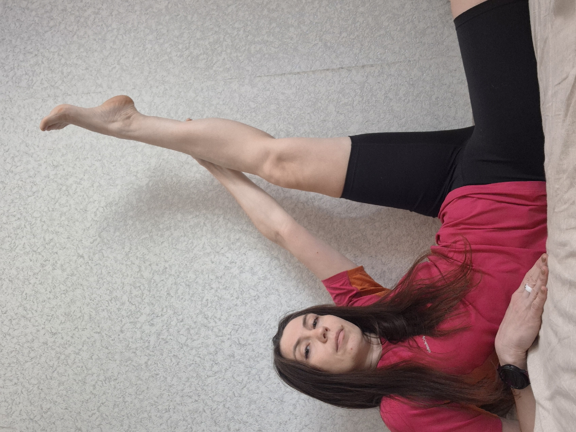 A woman is lying on her side on a bed, with her head towards the bottom of the image and her feet towards the top. She is wearing a red t-shirt and black leggings. Her right leg is extended upward, perpendicular to her body, while her left leg is bent. Her right arm is extended upward, and her left arm rests on the bed. She has long brown hair and is looking directly at the camera. A ring is on her left hand, and a watch is on her left wrist. The background is a white wall with a speckled texture. The location is Unknown, Unknown.