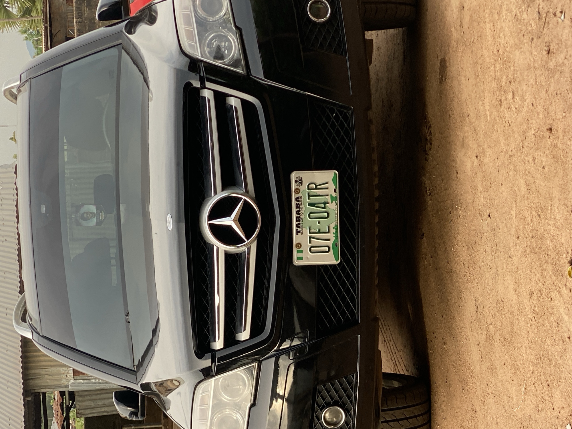 The image shows the front of a black Mercedes-Benz SUV parked on a dirt ground. The car's license plate reads "07E-04TR" and indicates "TARABA" and "FEDERAL REPUBLIC OF NIGERIA." In the background, there is a building with a corrugated metal roof and some vegetation. The environment appears to be an outdoor setting, possibly in Jalingo, Nigeria, given the license plate. A small picture of a man is visible on the interior of the windshield, to the left of the steering wheel.