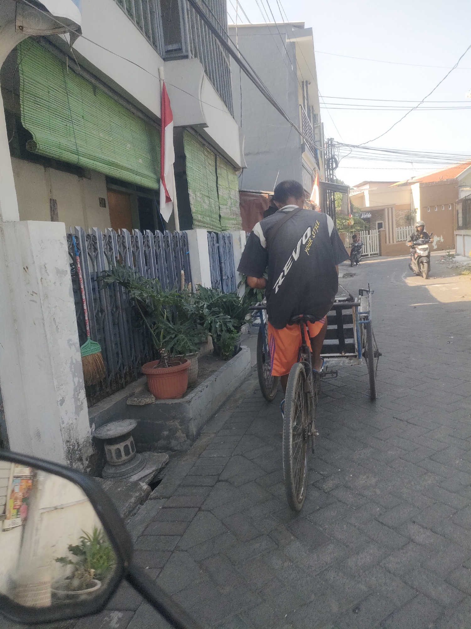 A man wearing a black shirt with white lettering that reads "REVO YOUR STYLE" and orange shorts is riding a three-wheeled bicycle on a paved street in Surabaya, Indonesia. The man is facing away from the camera and appears to be transporting something in the cart behind him. The street is lined with buildings and fences, and there are potted plants along the side of the road. A motorcycle is visible in the background. The image was taken during the day, with the sun casting shadows on the street.