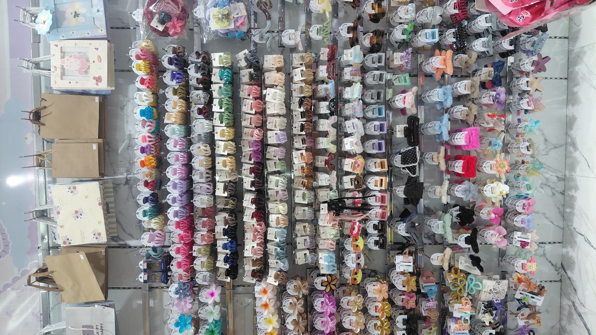 The image shows a retail store display of hair accessories, primarily hair clips and claws, organized on metal shelves. The items are brightly colored and come in various shapes, including flowers, stars, and geometric designs. Some are solid colors, while others have patterns or fuzzy textures. The accessories are individually packaged on white backing cards with product information. Above the hair accessories, there are displays of gift bags and decorative items, including paper bags and framed pictures. The overall environment appears to be a well-lit store with a white marble-patterned wall in the background and a light-colored tiled floor. The image does not provide any specific location information within Purwokerto, Indonesia. There are no people visible in the image.