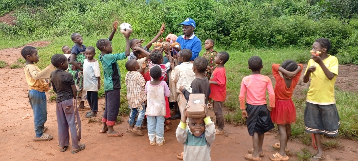In Ngalula, Democratic Republic of the Congo, an outdoor scene depicts approximately 20-25 children gathered around a single adult male on a reddish-brown dirt path. The background features dense green foliage, indicative of a natural or undeveloped area.
The adult male, positioned centrally, wears a blue long-sleeved shirt and a blue baseball cap with a white emblem. He holds a light-colored basket containing numerous irregular-shaped items, potentially food or small objects, which he is presenting or distributing to the surrounding children.
The children, ranging in age from toddlers to pre-teens, are dressed in various casual clothing including t-shirts, shirts, shorts, and skirts; many garments exhibit visible dirt. Their footwear consists of sandals, crocs, or bare feet. Several children are clustered around the adult, with some extending their arms towards the basket. A child on the left side of the frame holds a black and white soccer ball overhead. In the immediate foreground, a young child faces the camera with a smile, balancing a small, light-brown, rectangular object on their head. On the far right, a girl in a yellow shirt is actively eating a light-colored, spherical fruit or snack, while another child beside her stands with hands clasped behind their head. The overall interaction suggests an organized distribution or sharing activity.
