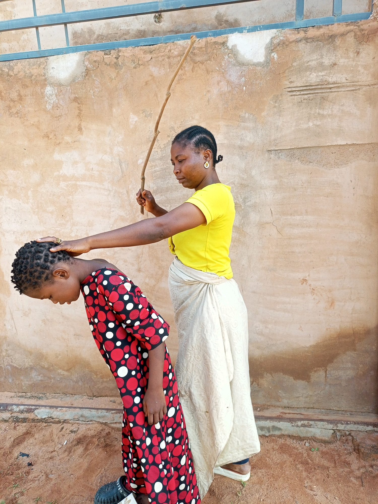 In Tempure, Nigeria, a woman wearing a yellow t-shirt and a light-colored wrap skirt holds a thin branch aloft, seemingly about to strike a child. The child, dressed in a red and black polka-dot dress, is bent over with their head down. The woman's left hand is on the child's head. They are standing outdoors against a rough, textured beige wall, with a metal bar visible at the top of the frame. The ground is composed of reddish-brown dirt and scattered small stones.