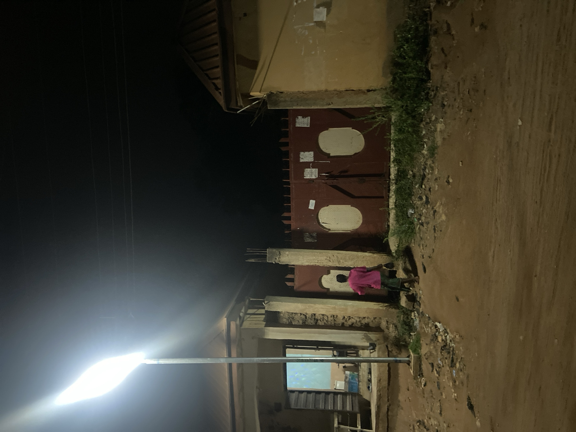 An image captured at night shows a street scene in Kuson, Nigeria. A person, seen from behind wearing a pink t-shirt and dark shorts, walks away from the camera towards a large, dark red gate. The gate has two oval-shaped indentations and is framed by concrete pillars. To the left of the gate, a building with an open doorway is illuminated from within. A street lamp with a bright, elongated light fixture stands tall on the left side of the frame, casting a strong glow on the surrounding area. The ground is unpaved and appears to be dirt, with scattered debris and vegetation visible along the edges of the scene. Power lines are visible against the dark night sky.