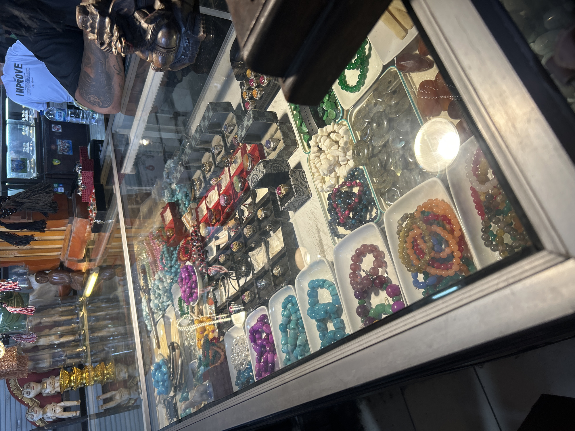 The image shows a close-up view of a glass display case filled with jewelry and decorative items. The display case is filled with bracelets made of various colored beads, including blue, purple, green, pink, and orange. There are also rings with colorful stones, and other small trinkets. In the background, a person wearing a white t-shirt with the word "IMPROVE" printed on it can be seen. The scene appears to be a stall or shop, possibly in a market. The lighting suggests it is indoors. The location is identified as Denpasar, Indonesia, likely a tourist area or a local market selling handicrafts and jewelry.