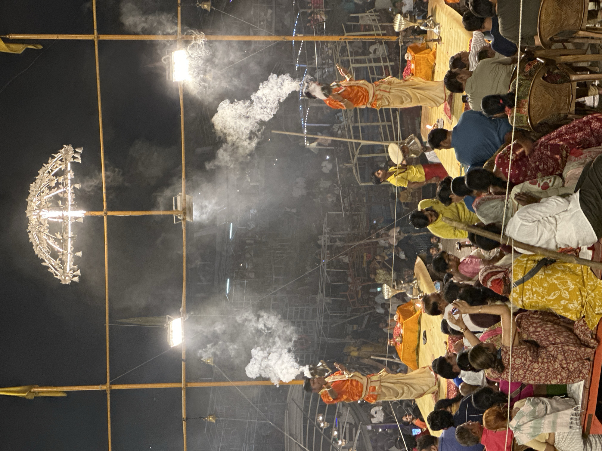 The image shows a night scene in Varanasi, India. A large crowd is gathered to watch a ritualistic fire ceremony. Priests dressed in orange and tan robes stand on platforms and direct billows of white smoke toward the crowd. Suspended above the scene are bamboo structures supporting spotlights and decorative hanging lights. The darkness of the night sky provides a backdrop for the illuminated spectacle.
