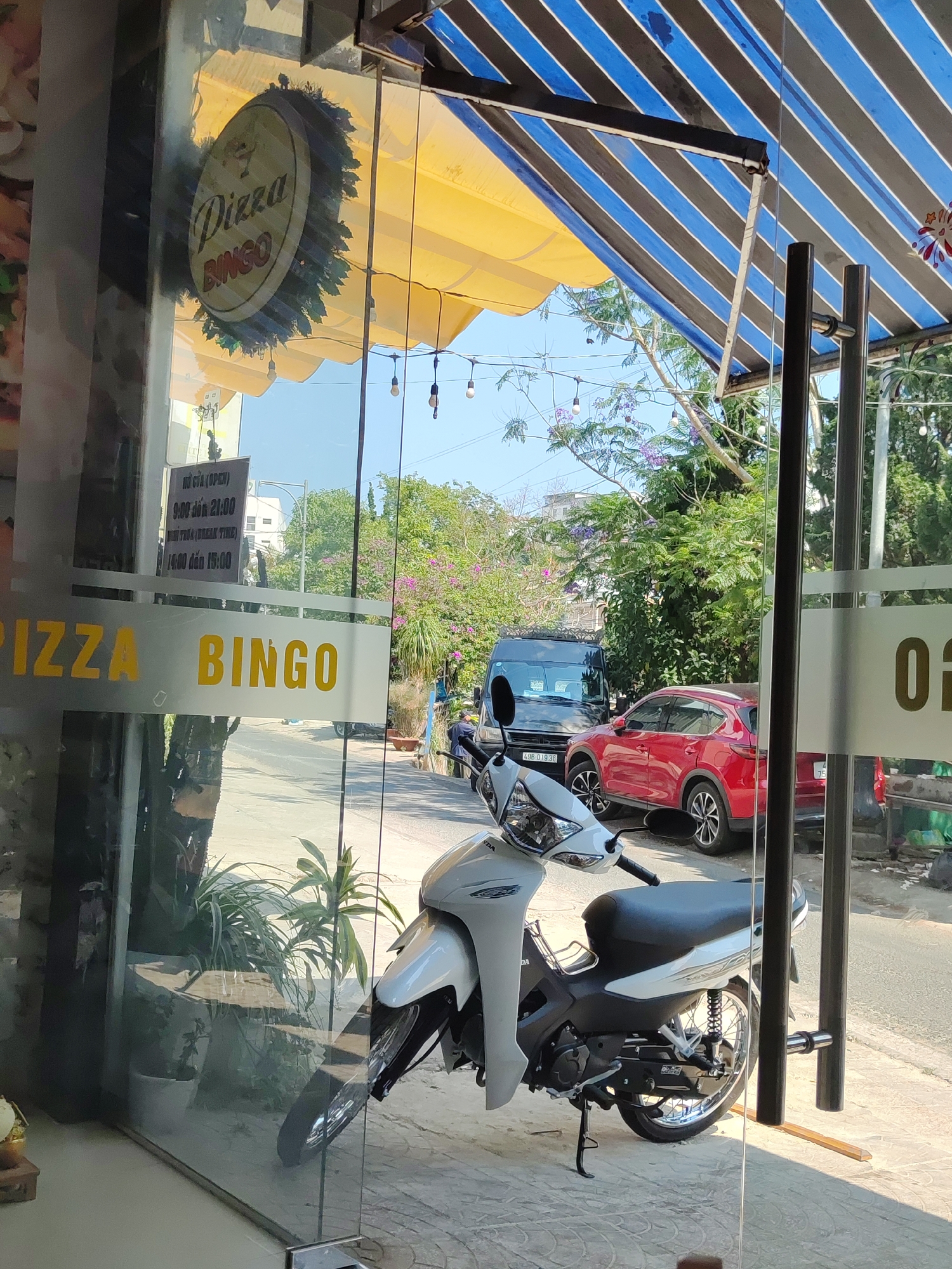 The image shows the front of "Pizza Bingo" in Da Lat, Vietnam. A white scooter is parked in front of the glass entrance doors. Through the glass, a sign indicates the business hours: 9:00 to 21:00, with a break time from 14:00 to 15:00. A red car and a black van are visible on the street outside. The storefront has a yellow awning and a blue-and-black striped canopy overhead. Trees with blooming purple flowers line the street.