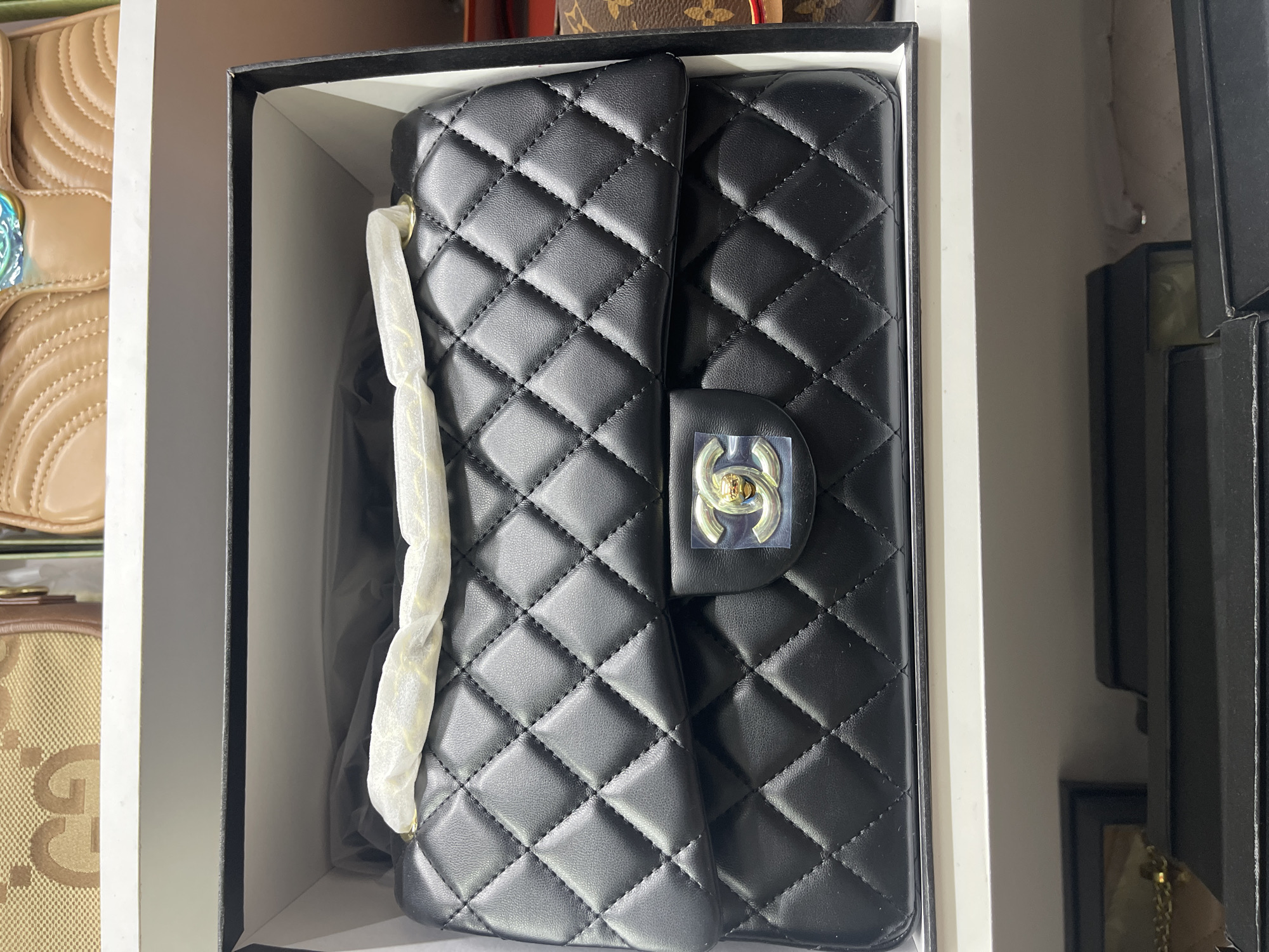 A black quilted leather handbag with a gold interlocking "C" logo is displayed in a black box. The handbag is new and wrapped in white tissue paper. The box is open, revealing the contents. In the background, other bags are visible on a white shelf. The image appears to be taken in a retail setting, possibly a store or a display area. There is no information available to suggest the location is Moscow, Russia.