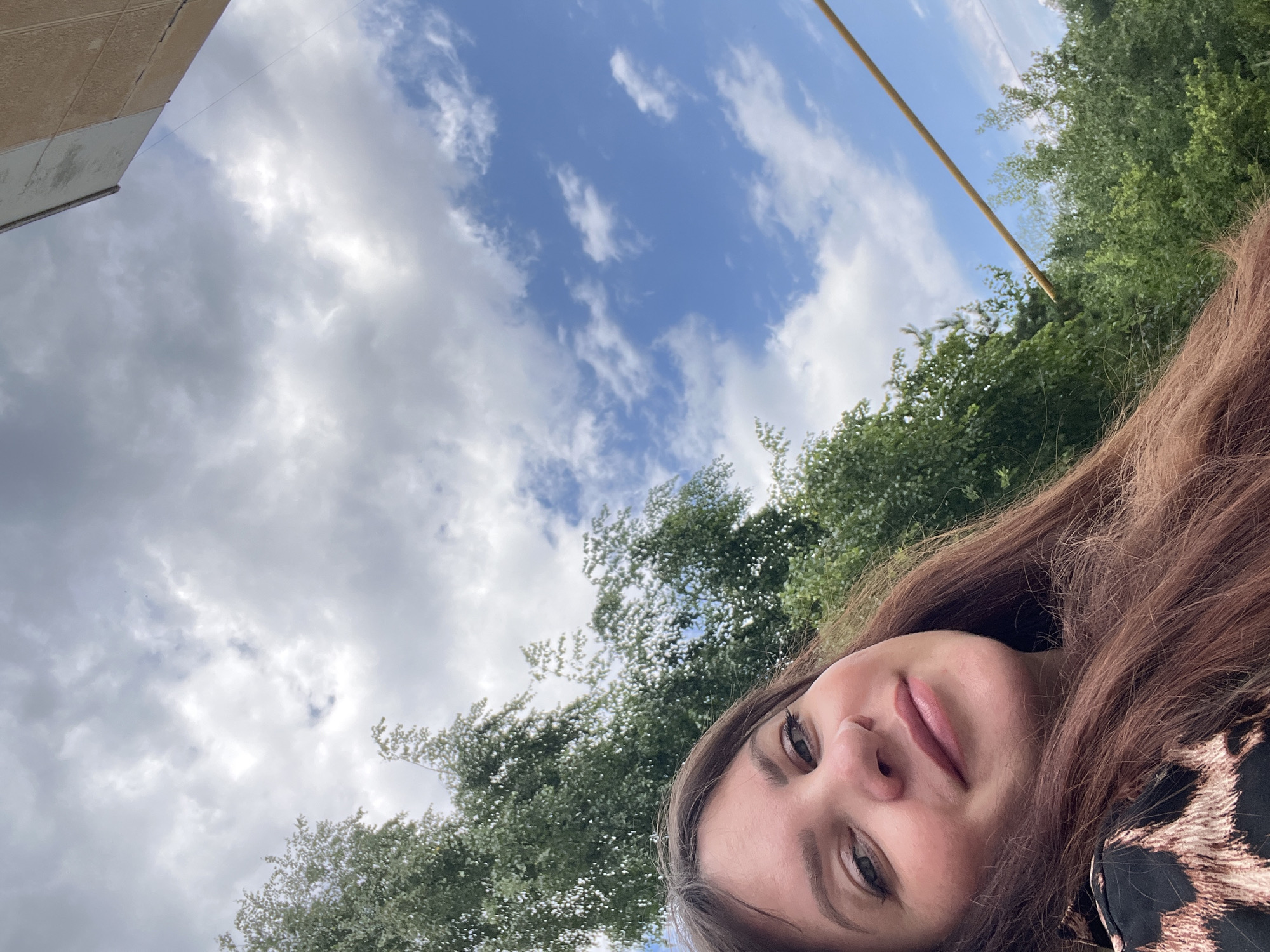 A woman with long, brown hair and light-colored eyes is captured in a close-up selfie outdoors. She is wearing a black top with a leopard print. Behind her, lush green trees and a blue sky with scattered white clouds are visible. A portion of a building, possibly an apartment building, can be seen in the upper right corner. The image appears to be taken in Noviy, Russia, judging by the presence of common Russian apartment buildings and the foliage typical of the region.