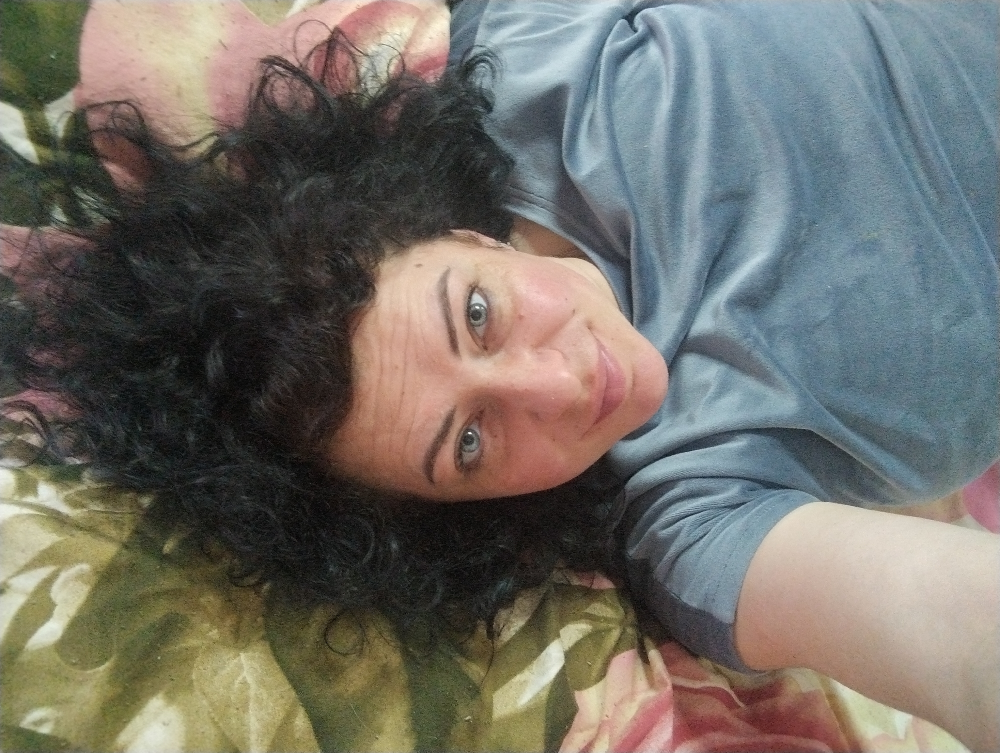 A woman with dark, curly hair is lying on a surface with a floral pattern. She is wearing a grey shirt with sleeves. Her eyes are light-colored. The image appears to be a close-up selfie. The location is not specified in the image and cannot be determined from it.