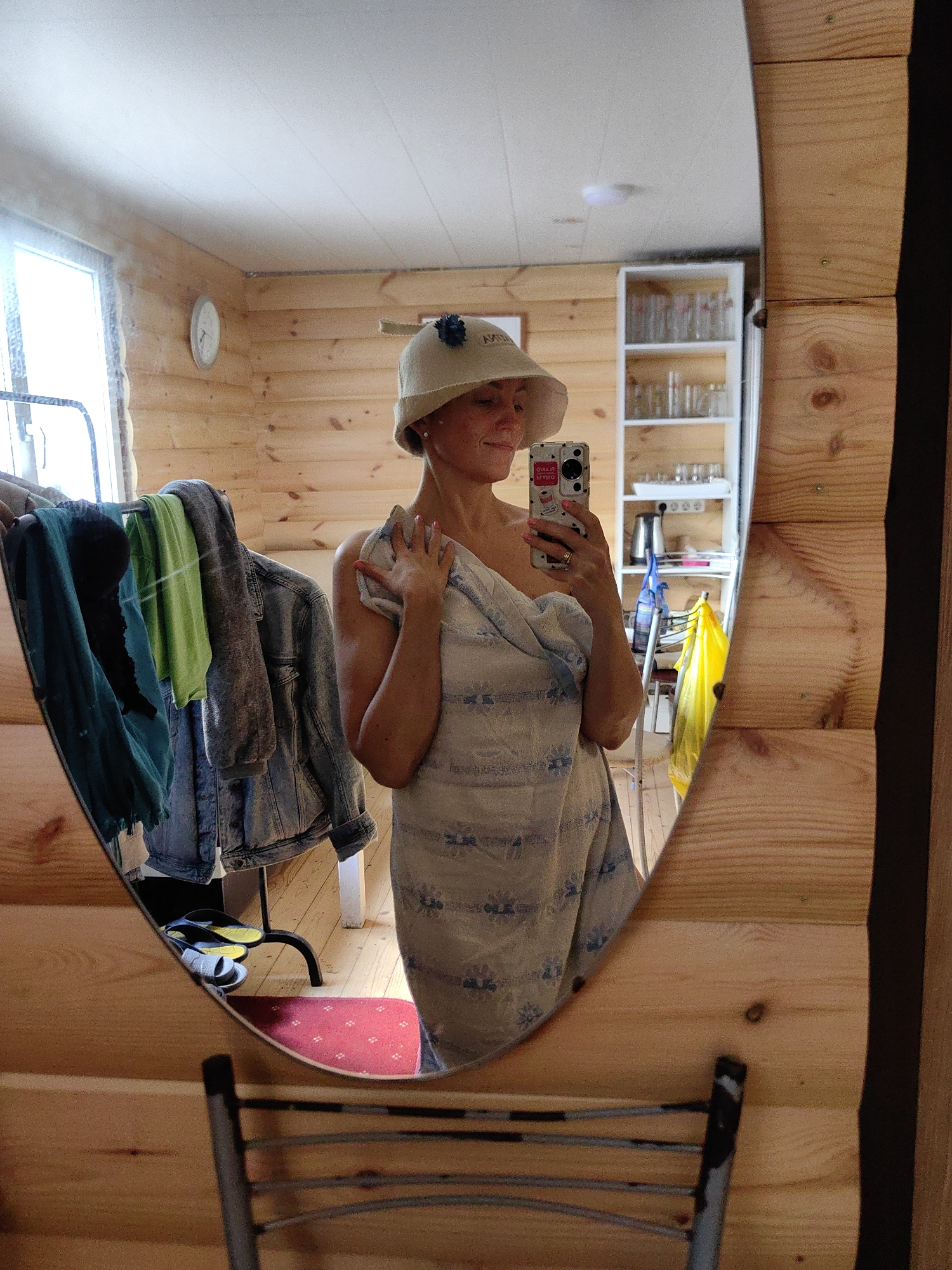 A woman is reflected in a large oval mirror, wearing a wide-brimmed hat and wrapped in a towel or blanket. She is holding a smartphone in her right hand, taking a selfie. The environment appears to be a wooden-paneled room, possibly a sauna or a changing room, with a window on the left and shelves filled with glasses in the background. Clothing is hanging on a rack to the left of the woman. The location is specified as Фаніпальскі сельскі Савет, Belarus.