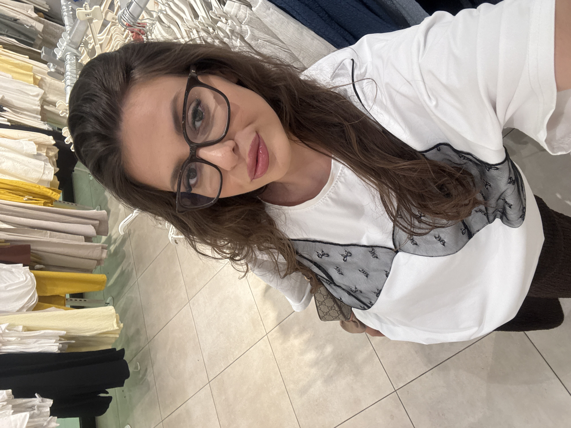 A woman with long, dark brown hair and glasses is taking a selfie in a clothing store. She is wearing a white top with black lace detailing and dark brown pants. She is holding a small purse with a monogrammed pattern. The background shows racks of clothing in various neutral tones. The floor is tiled. The location is a retail environment.