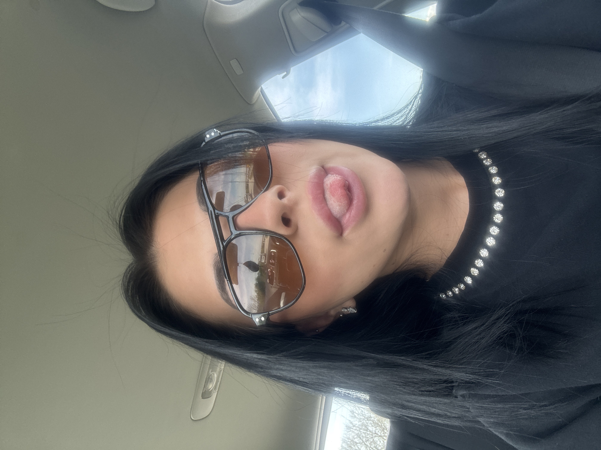 The image captures a close-up, angled shot of a young woman inside a vehicle, likely a car, in Tsaratsovo, Bulgaria. She is partially visible, focusing on her upper face, neck, and shoulder. She wears large, aviator-style sunglasses with dark frames and amber-tinted lenses, through which reflections of the outside world, including trees and sky, are visible. Her dark hair cascades down her shoulder. She is wearing a black top or hoodie adorned with a single, curved line of small, sparkling embellishments, resembling rhinestones or sequins, around the neckline or shoulder area. The woman is making a playful expression, sticking her tongue out slightly.
The setting appears to be a car interior, with a light gray or beige headliner and an overhead light visible in the background. A car door frame and possibly a seatbelt are also discernible. Through the car windows, a bright, clear to partly cloudy sky is visible, along with the silhouette of bare tree branches, suggesting it is daytime, possibly in spring or late autumn. The overall mood conveyed is casual and lighthearted. No visible text is present in the image.