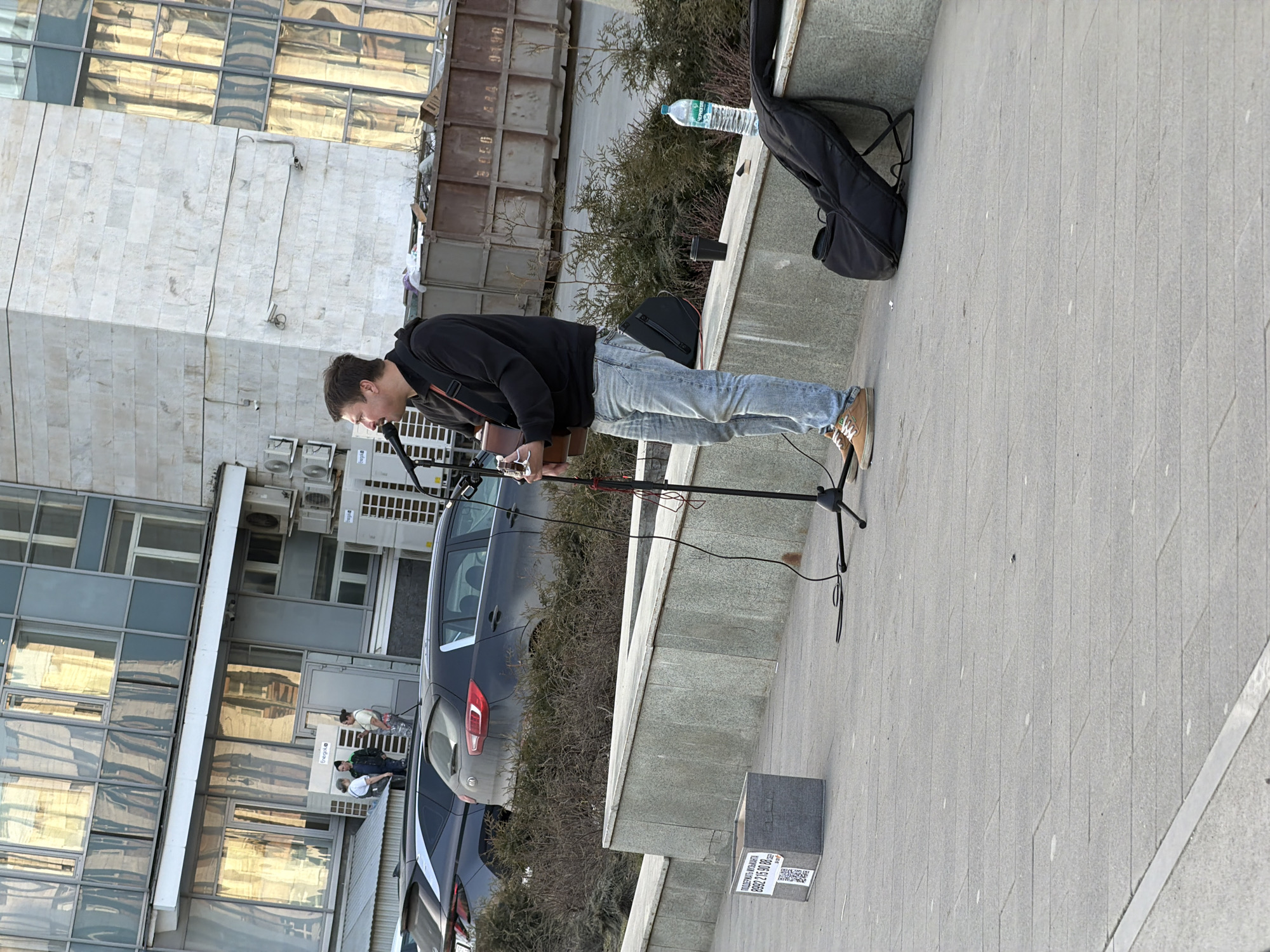 A street scene in Perm, Russia, shows a light-skinned male busker with short brown hair wearing a black hooded sweatshirt, light blue jeans, and brown shoes, playing a guitar and singing into a microphone. He stands on a grey brick pavement near a low concrete wall with bushes growing behind it. A black soft-sided guitar case and a plastic water bottle sit on the wall behind him. A grey hatchback vehicle is parked behind the wall. In the background, a modern building with mirrored windows reflects the sky and nearby structures, while another building with a gray metal exterior is visible to the right. A small box with a QR code and text is placed on the pavement, likely for donations. Several people are seen indistinctly near the building in the background.