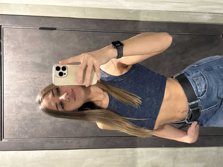 A young adult female is depicted in a mirror selfie, seen from the chest up to the upper thighs. She has long, straight, light brown hair and a faint smile. Her left ear displays a small earring. She is wearing a dark blue, ribbed, cropped tank top, revealing her midriff, and blue denim jeans secured with a black leather belt and a silver rectangular buckle. Her left hand rests on the waistband of her jeans near the buckle. She holds a light-gold colored smartphone with a clear case in her right hand, aimed at her reflection. A dark-colored smartwatch with a patterned band is visible on her right wrist. The background, reflected in the mirror, consists of a textured, dark gray panel or door on the left, featuring a vertical seam and a dark hinge, and a lighter, plain gray-beige wall section on the right. The specific location within Saint Petersburg, Russia, is not discernible from the provided image, which appears to be an indoor setting such as a fitting room or private residence.