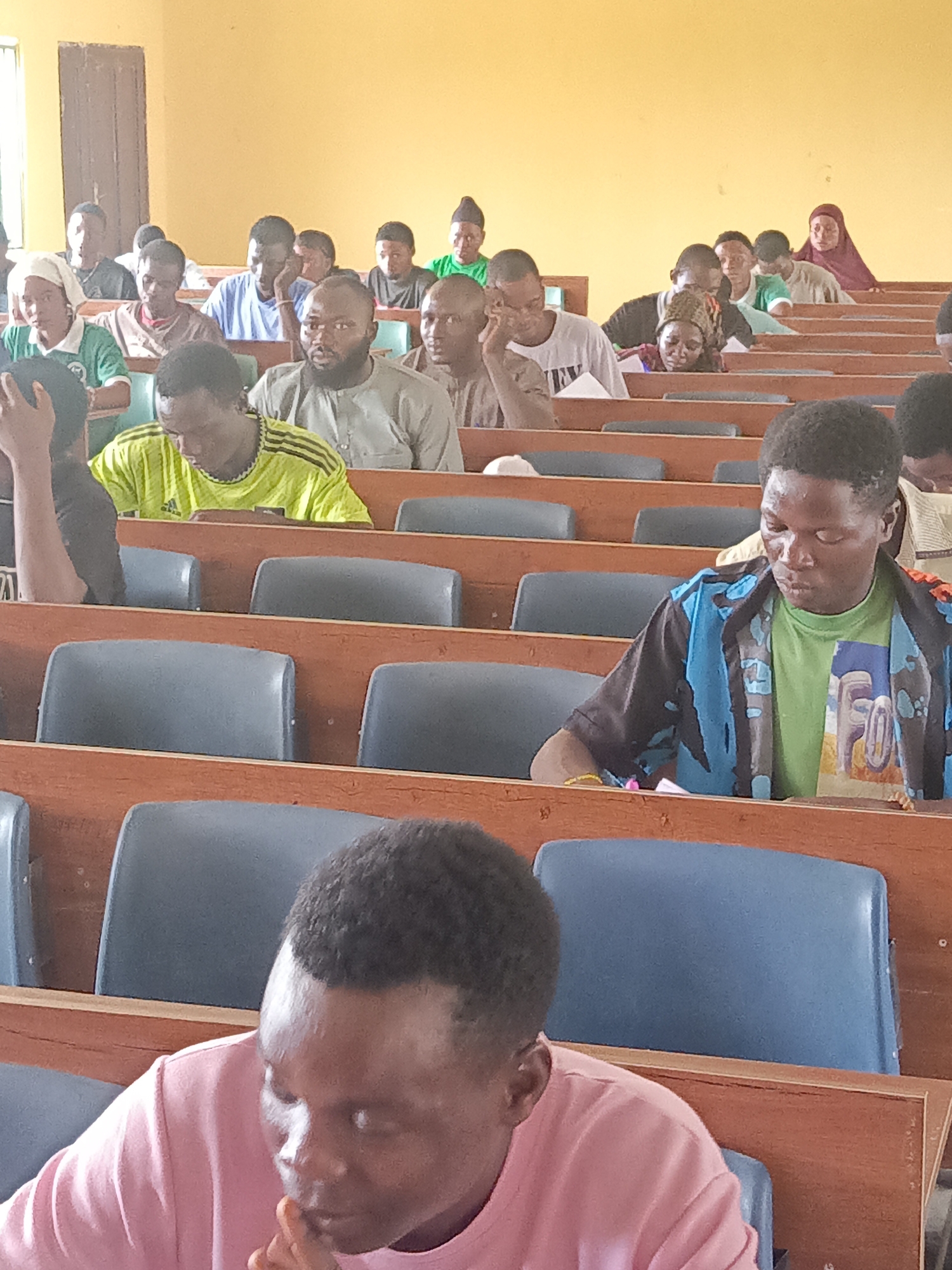This image captures a group of people, primarily young adults, attending a session in what appears to be a lecture hall or classroom in Akwanga, Nigeria. The setting features multiple rows of wooden desks with attached blue or grey seating, typical of an educational institution. The room has light yellow-painted walls, and natural light enters from a window visible on the left side, indicating it is daytime.
Approximately two dozen individuals are visible, occupying several rows of seats. Most are looking forward, suggesting they are listening to a speaker or instructor. Some individuals are looking down, possibly taking notes or reflecting. Their expressions are generally subdued, indicating attentiveness or contemplation; some have their hands resting on their chins. The attendees include both men and women, with some women wearing head coverings, such as a maroon hijab in the background and a white headscarf in the middle rows.
The attire is varied and casual, including t-shirts and collared shirts. One person in a bright yellow-green shirt displays what appears to be an Adidas logo on their chest. Another individual in the middle-right foreground wears a blue and black patterned shirt over a light green t-shirt with a graphic design that is not clearly legible. The overall activity depicted is a quiet gathering, likely a class, lecture, or meeting.