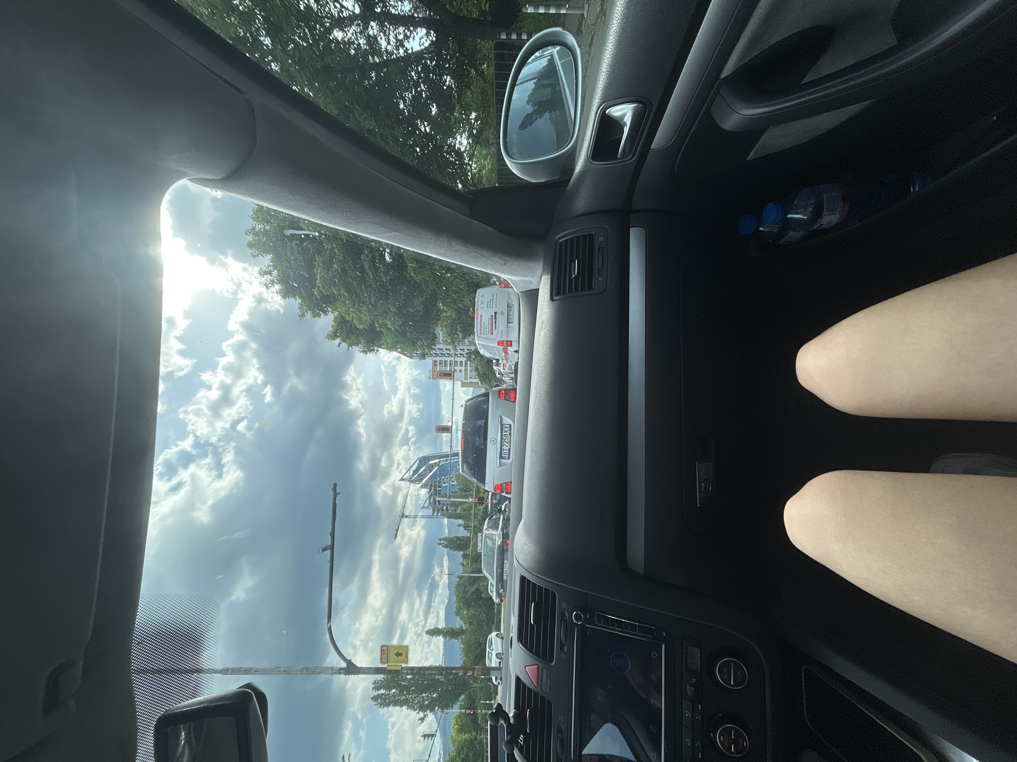 The image, taken from the driver's seat of a car, shows a view of traffic in Sofia, Bulgaria. The car is stopped in traffic, with other vehicles visible ahead. Construction cranes and building structures are visible in the distance, along with trees and a cloudy sky. A "BILLA" sign is also visible, suggesting the location is near a Billa supermarket. In the car's interior, part of the dashboard, a side mirror, and two water bottles in the door are visible. Two thighs of a person are visible to the right of the central console.