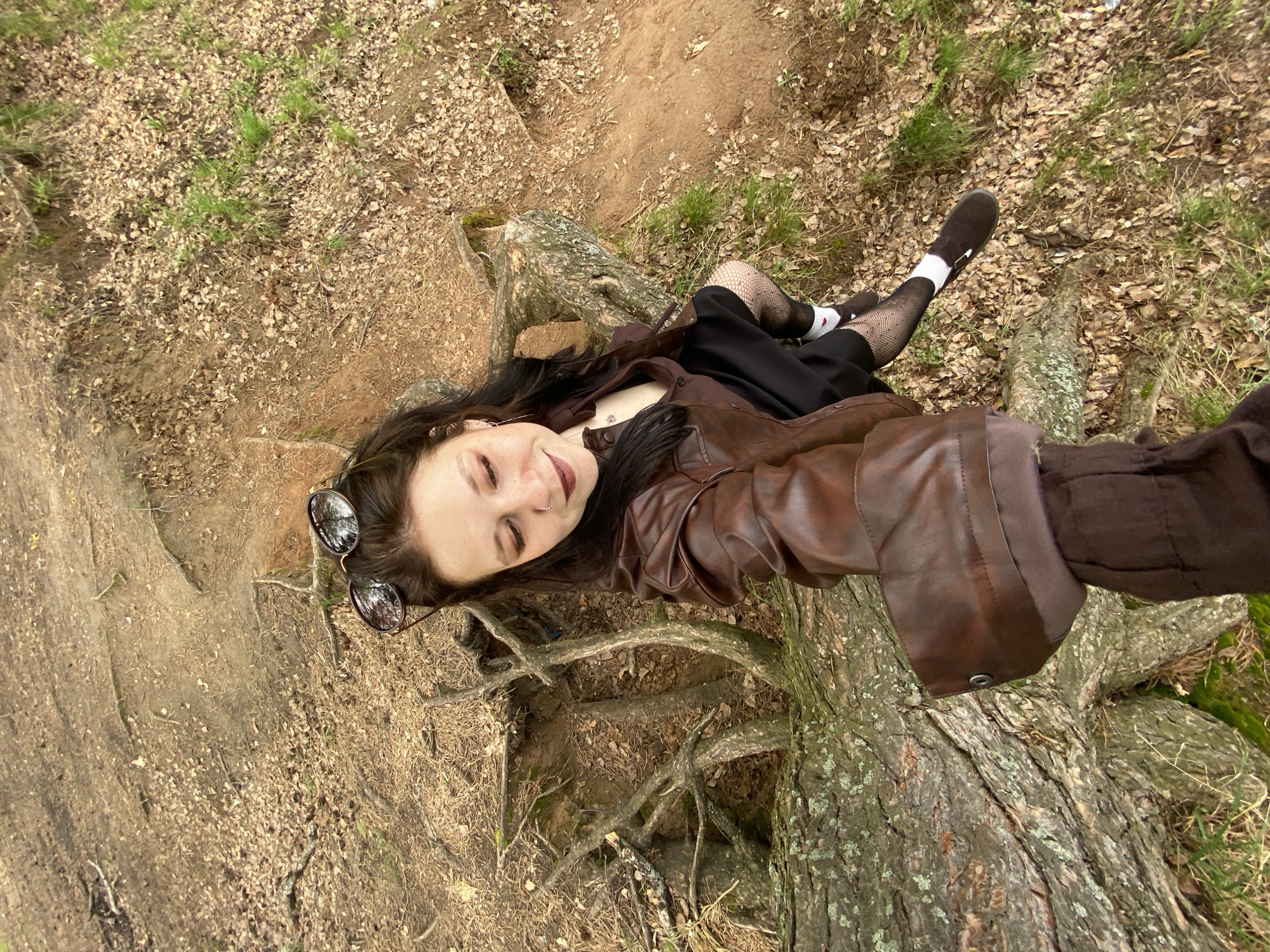 A woman is lying on a tree stump in an outdoor environment. She is wearing a brown leather jacket, a black dress, fishnet stockings, white socks, and black shoes. Sunglasses are resting on her head. The background consists of dirt, grass, and fallen leaves. The location appears to be a natural area, possibly in the city of Chaikovsky, Russia.