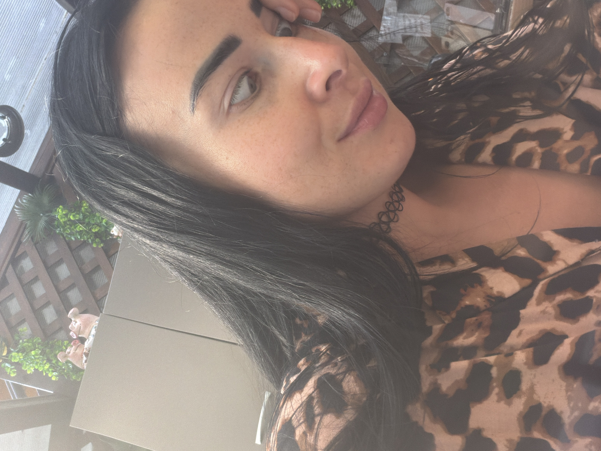 A medium shot shows a woman with long, dark hair and fair skin, looking downwards and to her right, with her right hand touching her face. She is wearing a leopard-print top and a black choker necklace. The background is out of focus, but appears to be an outdoor setting with a wooden lattice structure and greenery. The image does not contain any information about the city of Sochi, Russia.