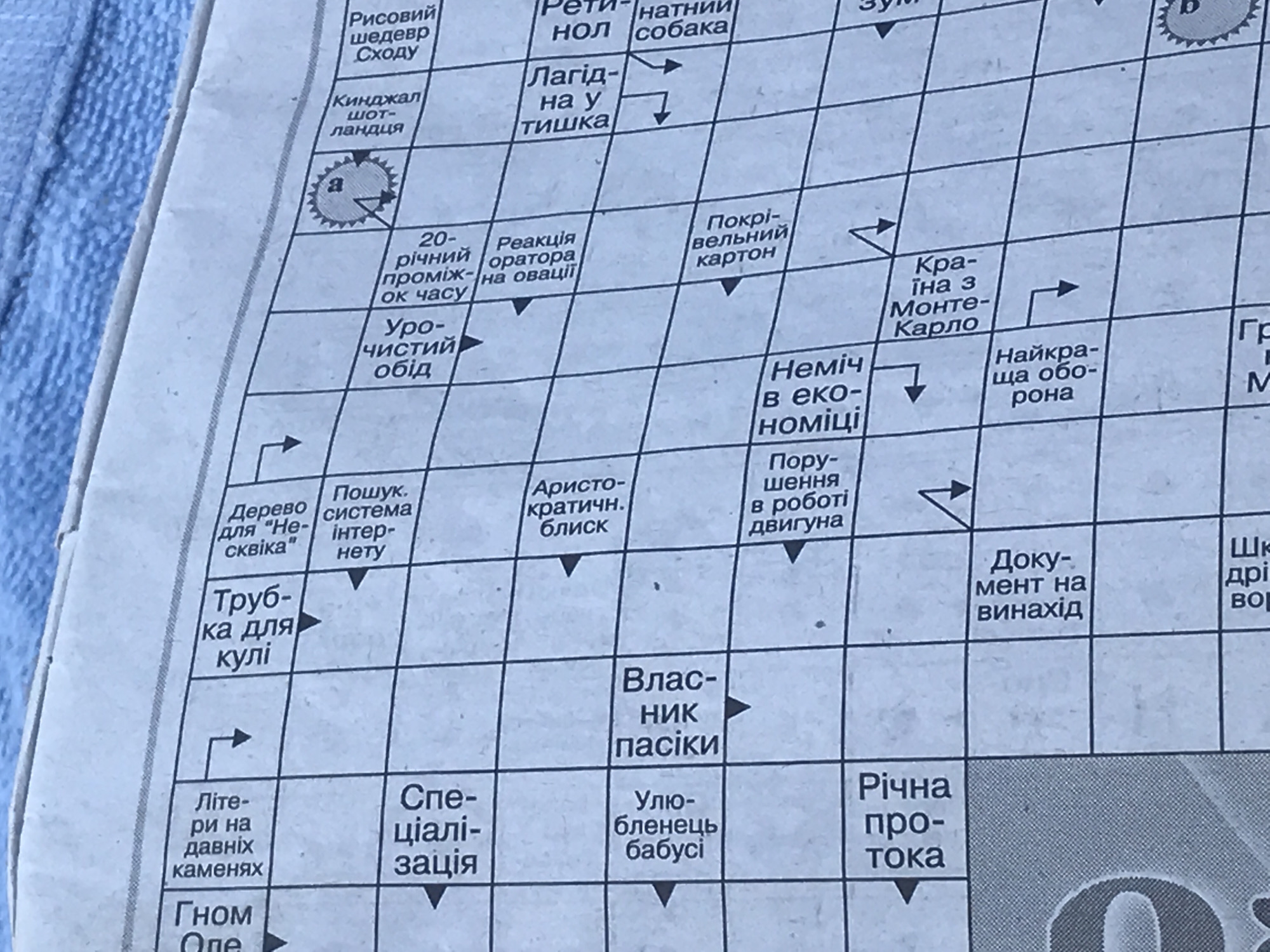 The image is a close-up of a section of a newspaper crossword puzzle, featuring Ukrainian text. The puzzle has a grid of cells with black lines separating them, and some cells contain words and directional arrows. The paper appears aged and slightly wrinkled. The background is a textured, light blue fabric, likely a towel or blanket. There are no people, animals, or any elements related to the city of Varna, Bulgaria.