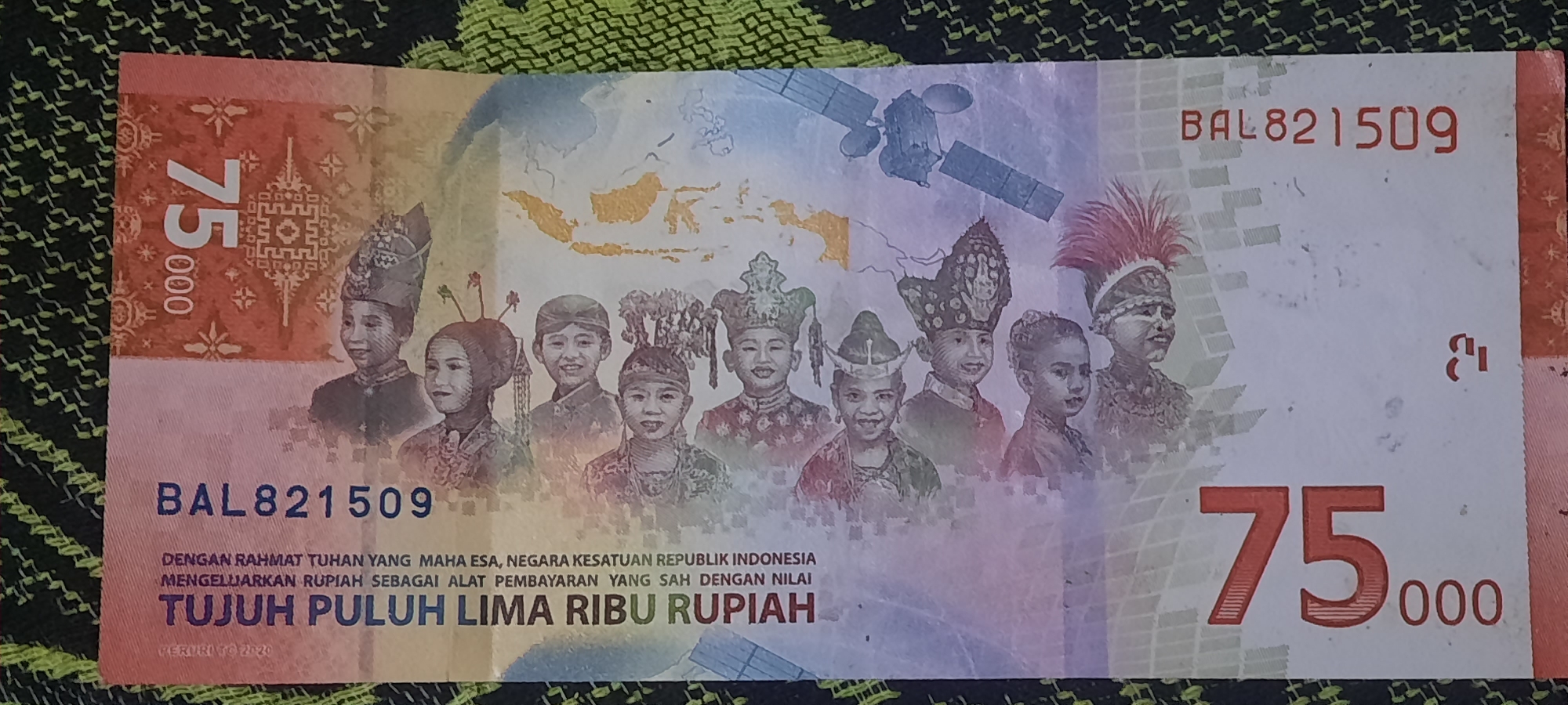 The image shows a close-up of an Indonesian banknote. The banknote is red and white and features illustrations of children in traditional attire, a satellite, and a map of Indonesia. The denomination "75.000" is prominently displayed. The text on the banknote indicates that it is legal tender of the Republic of Indonesia. The banknote is lying on a textured green and black surface. There is no visible indication of the location being the city of Jatitengah, Indonesia.