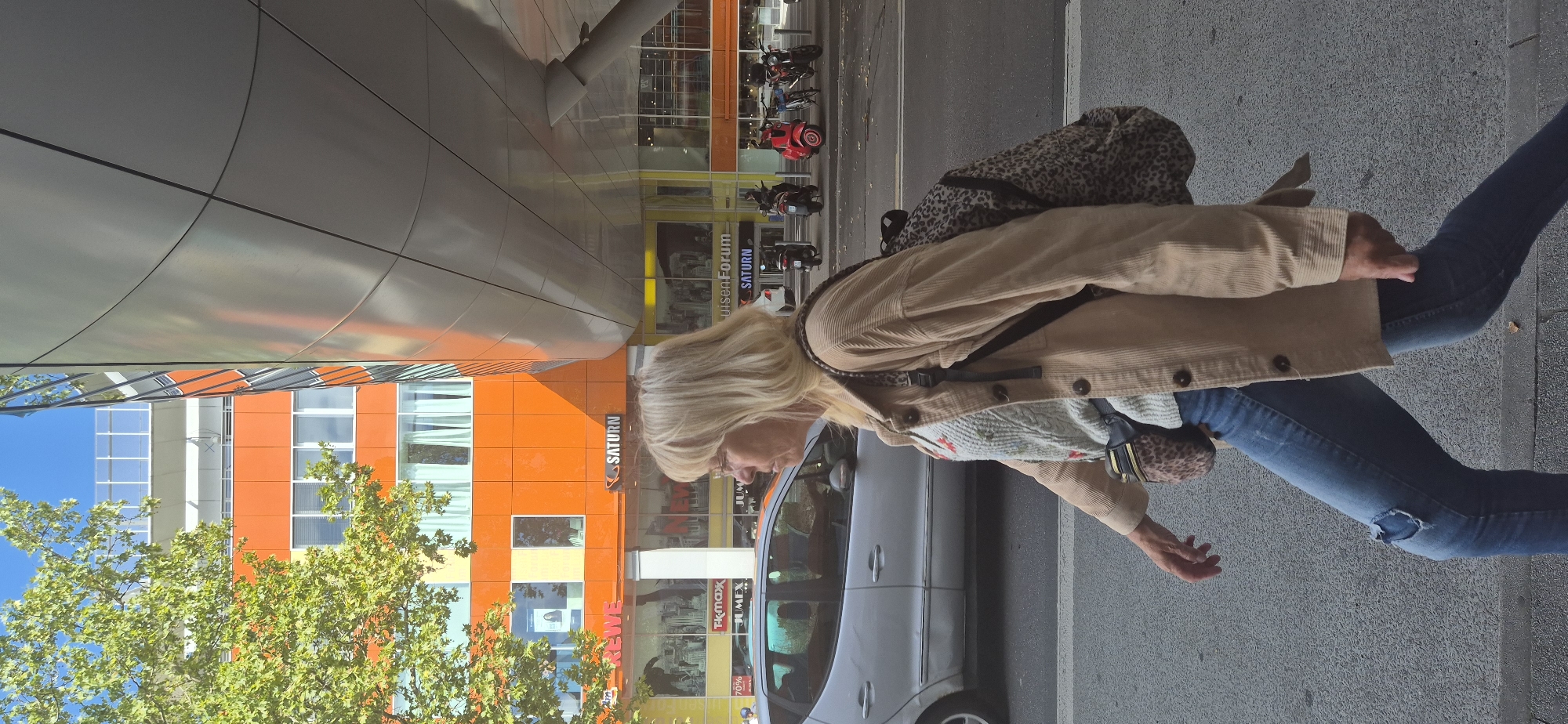 A middle-aged woman with blonde hair, wearing glasses, a light-colored corduroy jacket over a patterned sweater, ripped blue jeans, and a leopard-print fanny pack, walks along a city sidewalk. She carries a leopard-print backpack. In the background, a modern building with orange and silver facade elements is visible. Signs for "Rewe" and "Saturn" are displayed on the building's storefront. Several parked motorcycles and scooters are also present on the street. A silver car is driving on the road. The scene appears to be in Wiesbaden, Germany, likely on a sunny day given the shadows and bright lighting.
