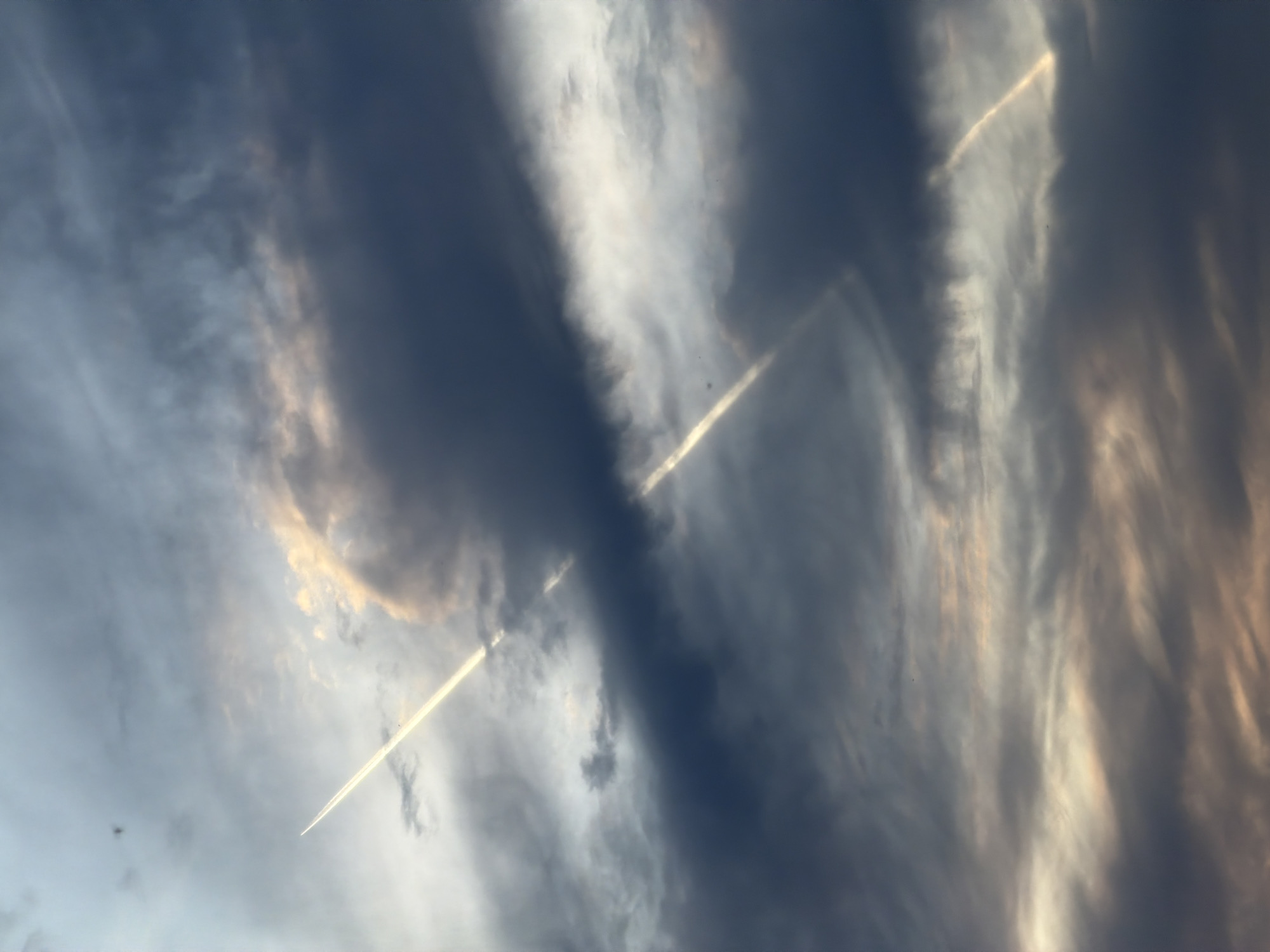 The image shows a cloudy sky with contrails. The clouds are a mix of dark and light shades, with some golden light appearing to shine through. Two contrails are visible, one running diagonally across the image from the lower left to the upper right, and another fainter one towards the upper right. There is no indication of the location or city of Chaikovsky, Russia.