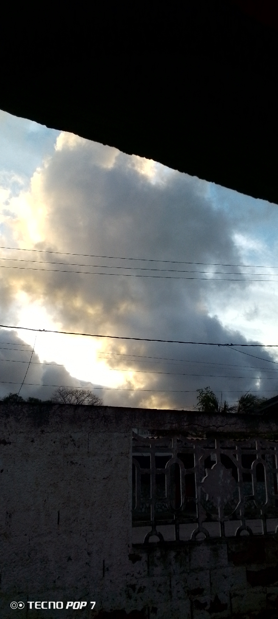 The image displays a dramatic sky scene, likely captured during either sunrise or sunset in Altagracia, Venezuela. Large, heavy clouds dominate the upper and middle sections, exhibiting a stark contrast between dark, dense formations and areas brilliantly illuminated by golden-yellow sunlight breaking through. Patches of lighter blue sky are visible, particularly towards the upper right.
Three dark utility wires stretch horizontally across the central part of the sky, with an additional diagonal wire visible on the far left. Below the horizon, a textured, light-colored concrete or stucco wall occupies the lower portion of the foreground. This wall appears weathered, with a visible crack along its top edge. On the right side of the wall, a dark, decorative metal or concrete grille featuring geometric patterns is integrated. Silhouetted against the bright sky are bare tree branches on the left, and some leafy green vegetation appears above the wall on the right. A dark, indistinct mass, likely a roof or overhang, partially frames the very top of the image.
There are no people or specific activities depicted. The scene conveys a moody yet striking atmosphere, characteristic of a dramatic sky over an urban or semi-urban setting. The bottom-left corner of the image contains the visible watermark "◎ TECNO POP 7", indicating the device used for the photograph.